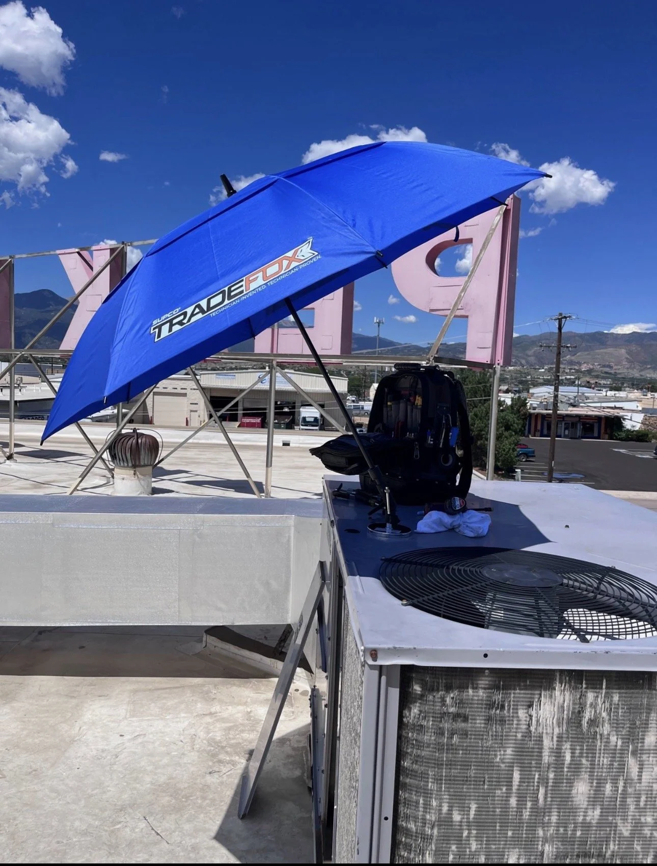 On a rooftop, there is a large blue umbrella with racing decals, a black backpack, and various tools on a white surface. In the background, a billboard, power lines, and mountains are visible under a blue sky with clouds.