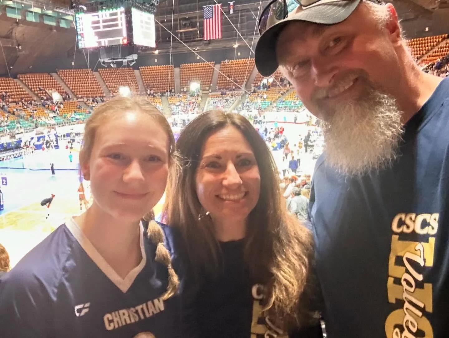 Three people smiling for a photo inside a sports arena with volleyball courts in the background. One young woman wearing a sports jersey on the left, a woman with long dark hair in the middle, and a man with a beard and cap on the right, wearing a shirt with text.