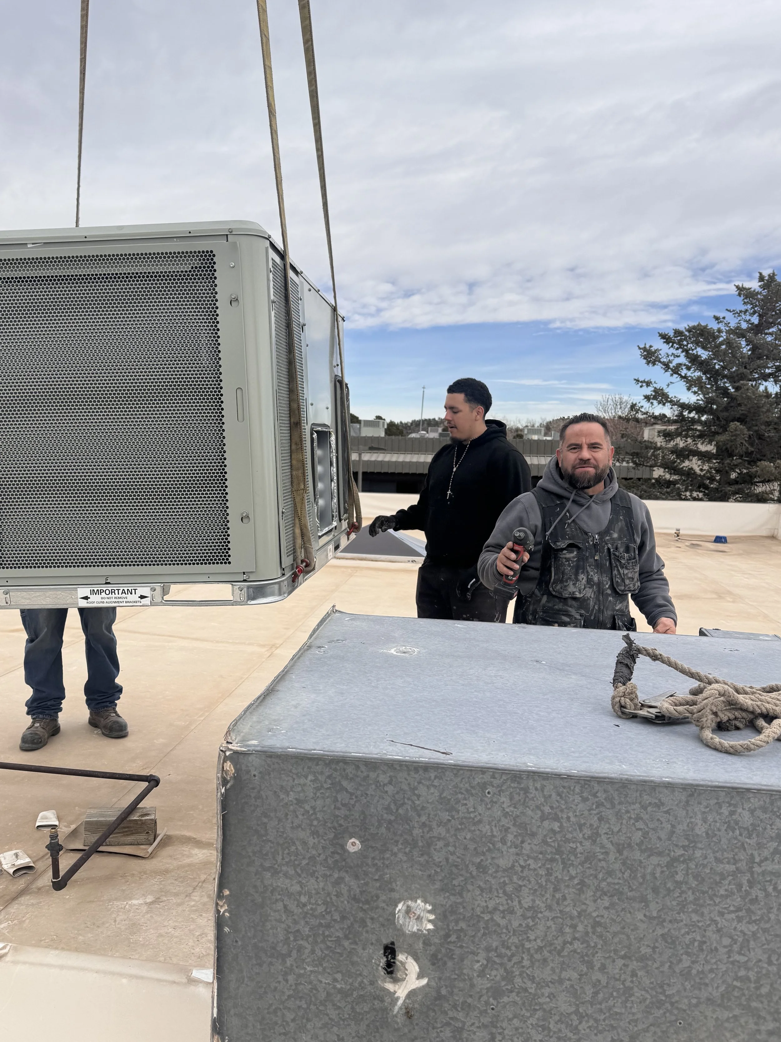 Two men working outdoors on a rooftop with large metal equipment, one is holding a power drill and the other is inspecting or adjusting equipment, with a cloudy sky and trees in the background.