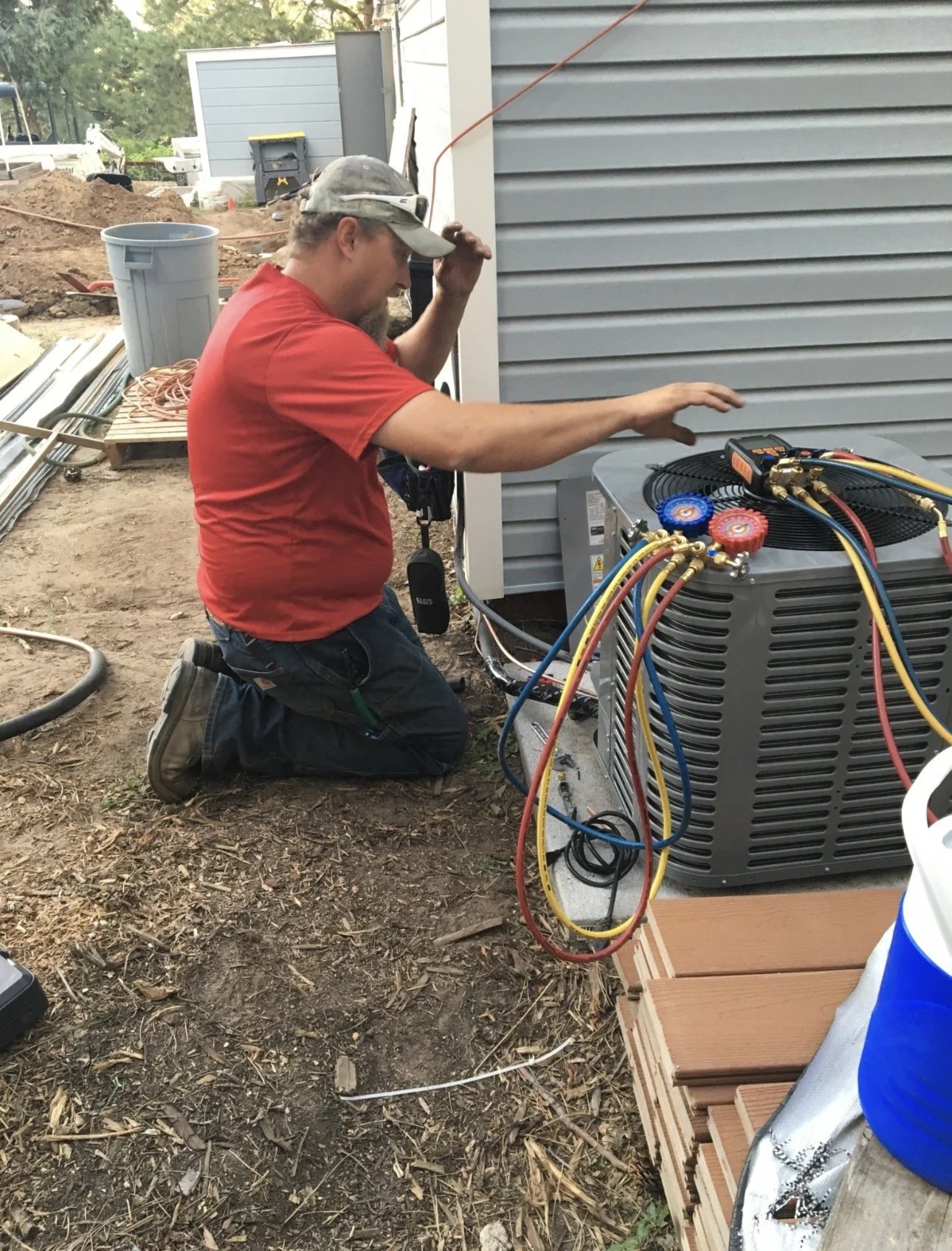 A man kneeling on the ground working on an HVAC unit outside a house. He is wearing a red shirt, jeans, a cap, and sunglasses, and is using tools connected to the unit with colorful hoses.