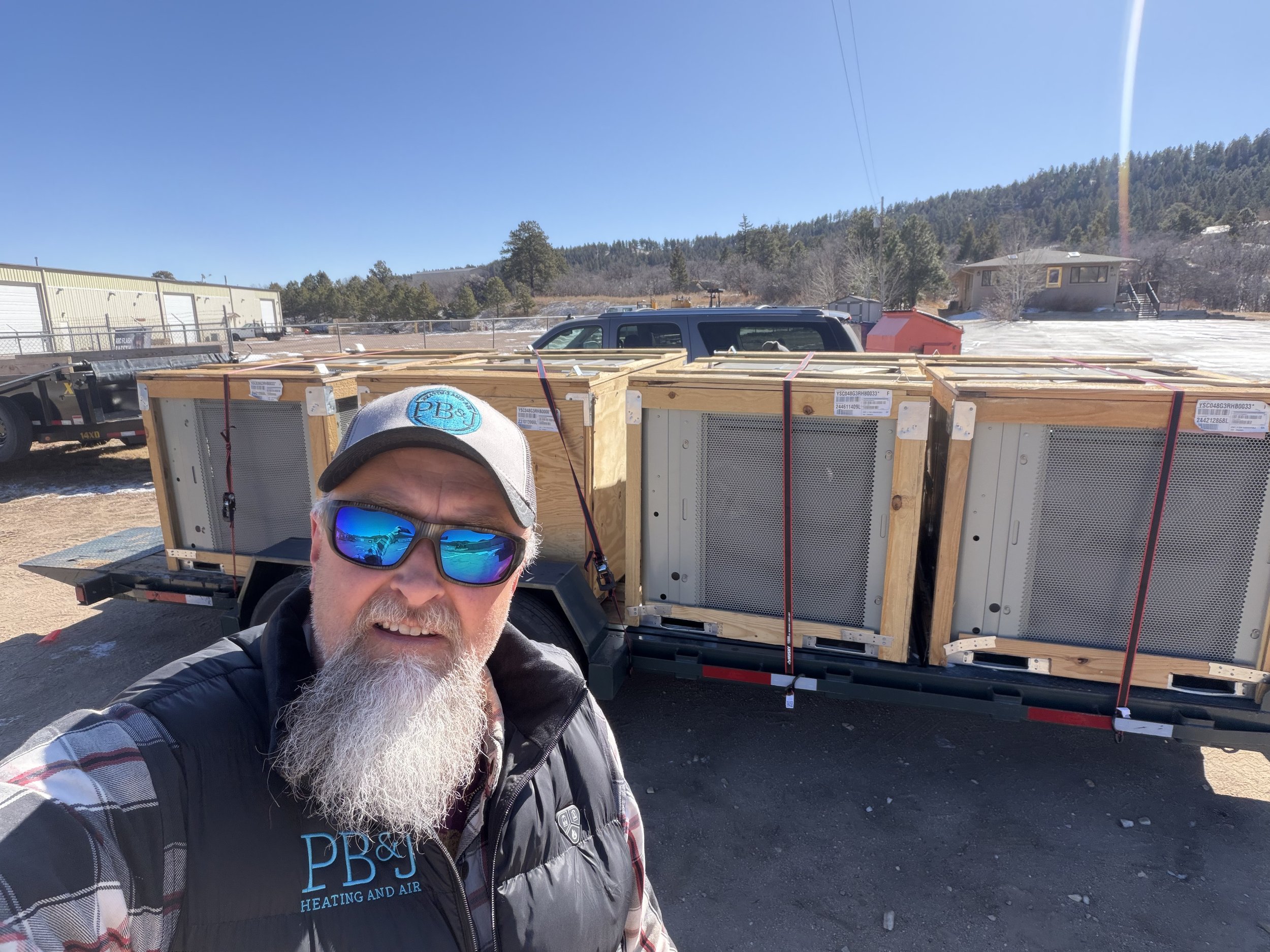 A man with a checked shirt, a black vest with blue logo, a white beard, and sunglasses taking a selfie outdoors near a trailer carrying large wooden crates, with a blue sky and a house on a hill in the background.