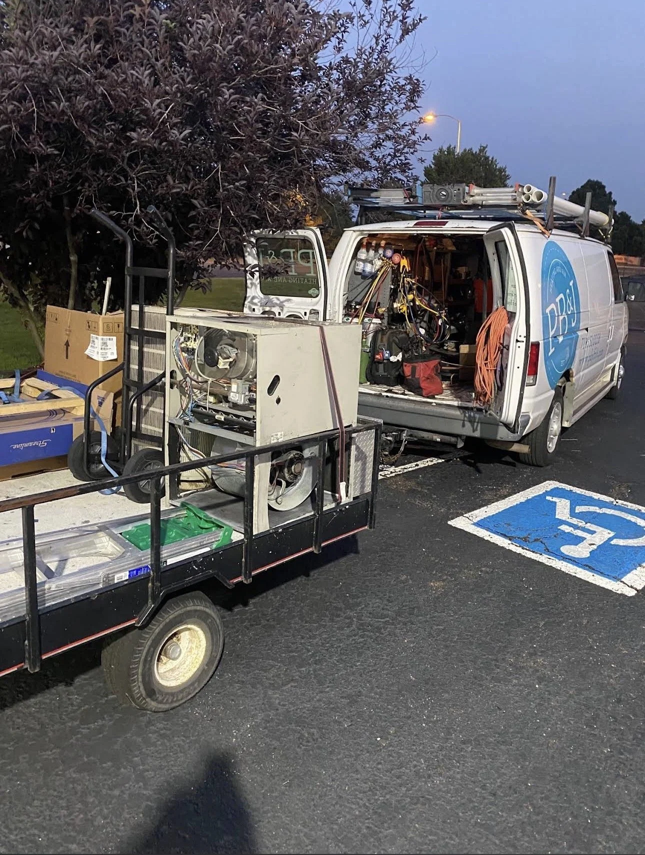 A white utility van with electronics equipment inside and on the roof, parked in a handicapped parking space next to a tree, with a utility cart loaded with electronic parts in the foreground.