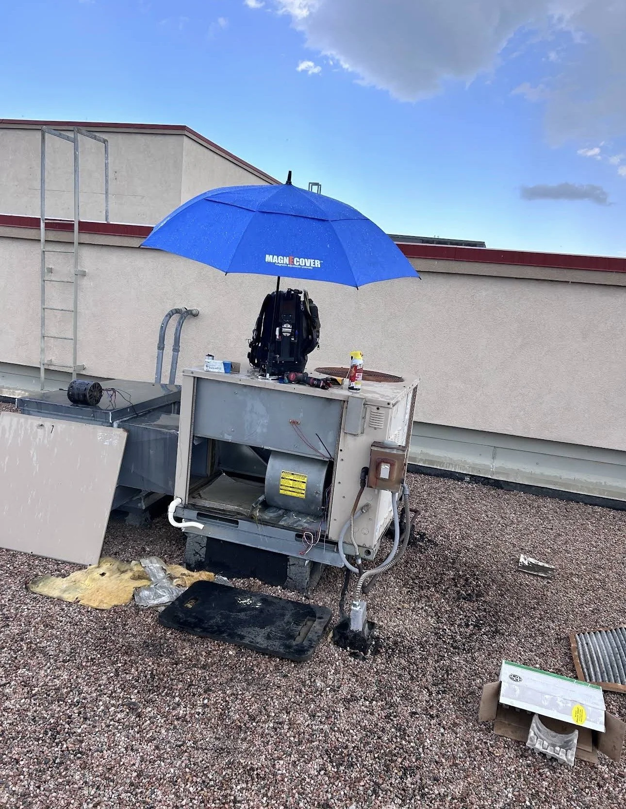 On a rooftop, there is an open HVAC unit with exposed components, surrounded by debris. A blue umbrella is attached to the unit, providing some cover. Various tools and personal items are placed on and near the unit. The background shows a beige buil