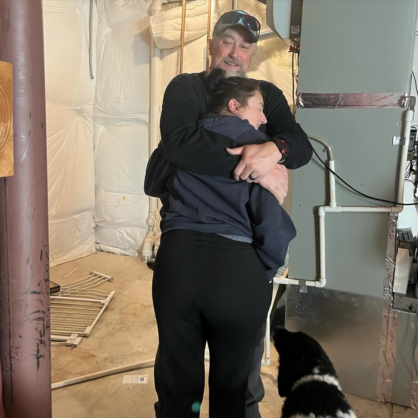 A man and a woman hugging in a basement or utility room. The woman is smiling with her eyes closed while the man looks to the side and is smiling. There is a black and white dog at their feet, looking up at them. The room has a white insulated wall, a boiler or furnace, and some plumbing pipes.