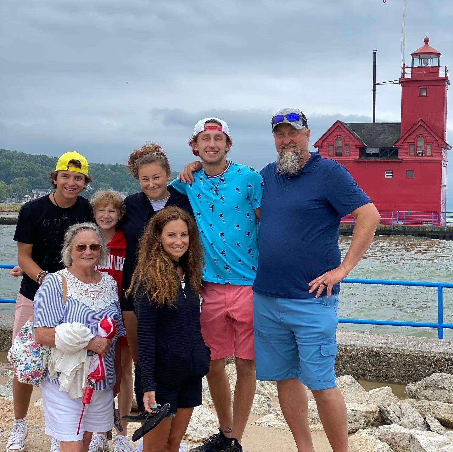 Group of eight people posing by the water with a red lighthouse in the background on a cloudy day.