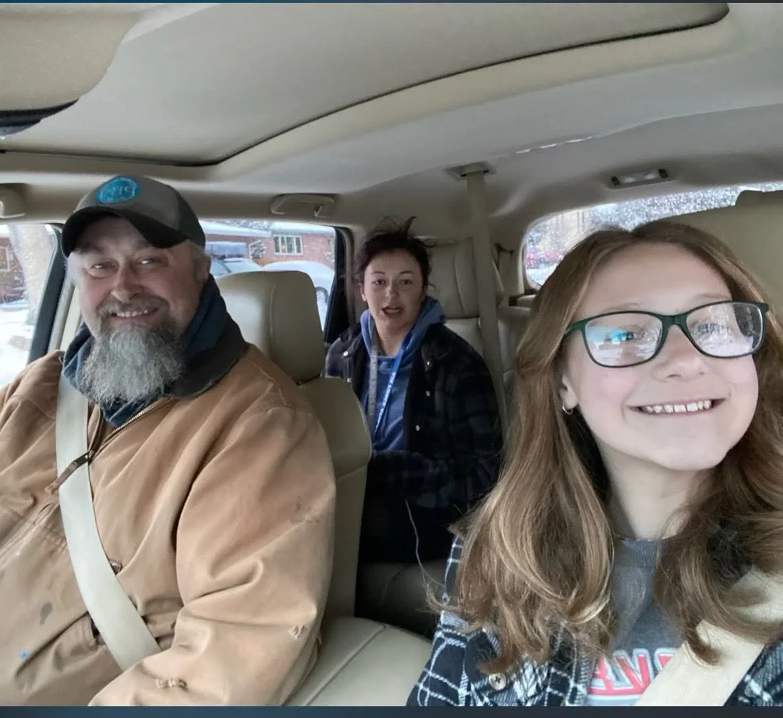 Three people sitting in the backseat of a vehicle, smiling. The person in front is a man with a gray beard and black cap, the middle person is a young woman with messy hair wearing a jacket, and the person on the right is a girl with glasses and long hair, taking a selfie.