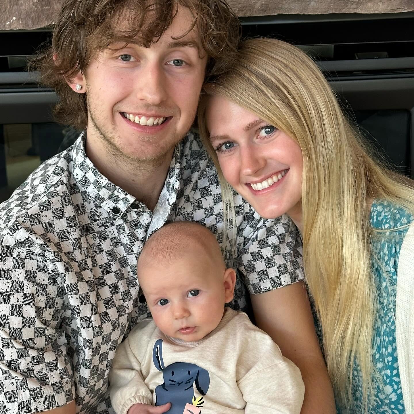 A smiling young man, a young woman, and a baby are close together, posing for a photo indoors.