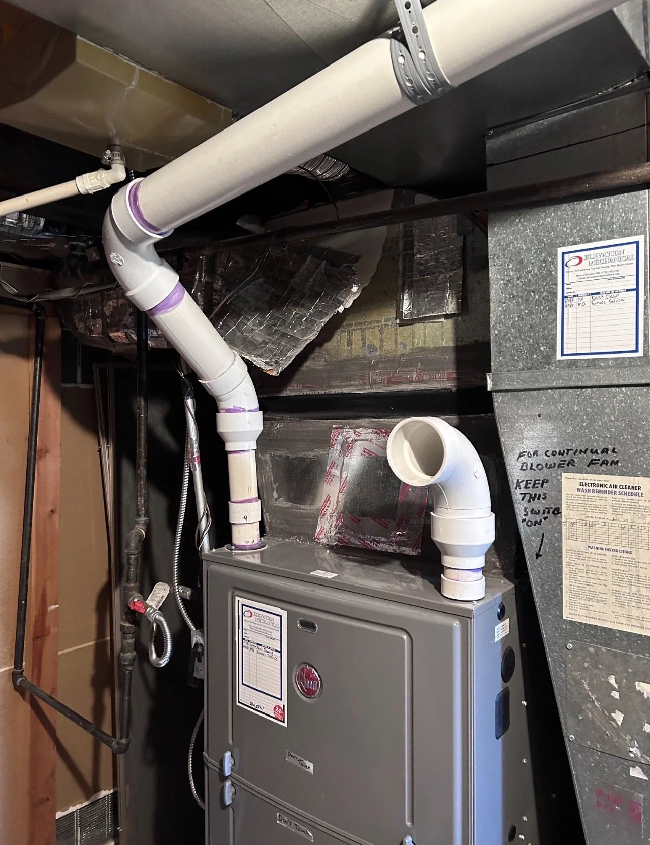 A furnace installed in a basement utility area with white PVC pipes connected to it and a metal duct. There are insulation materials and a schedule sheet attached to the ductwork. The furnace is gray with control labels on the front.