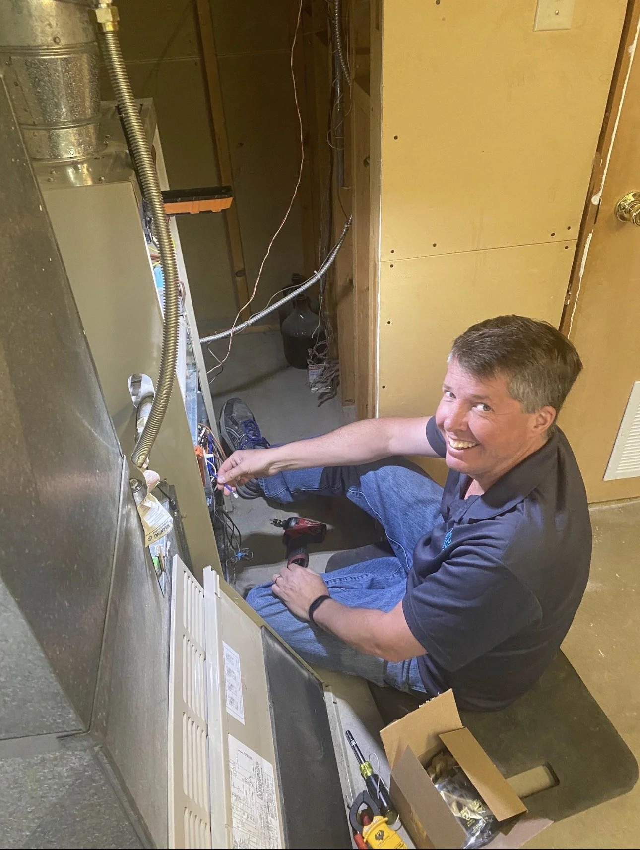 A man working on electrical wiring inside an unfinished room, smiling at the camera, using tools and surrounded by electrical components and wires.