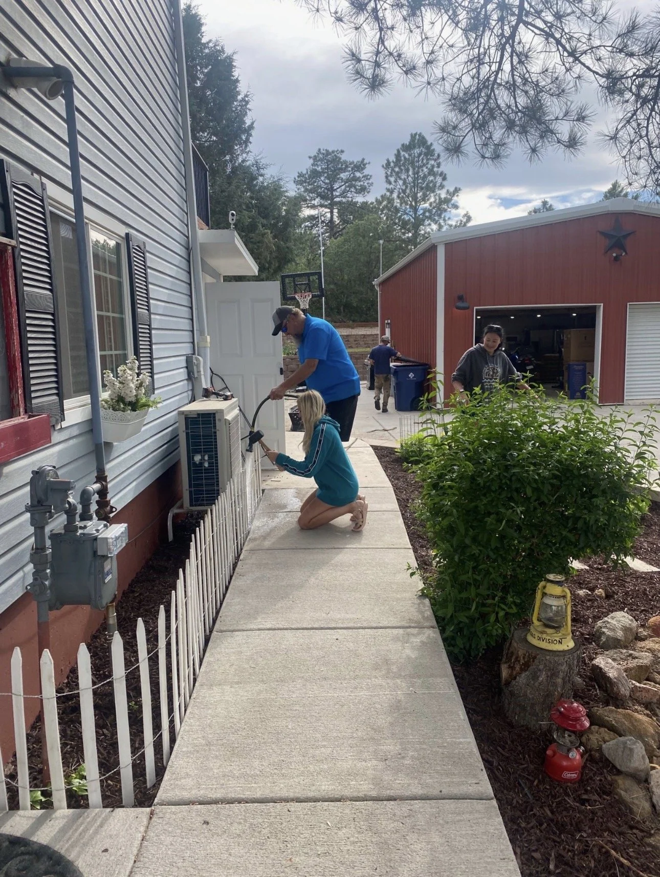A family of four is cleaning the exterior air conditioning unit outside a house. The father and daughter are kneeling and working together on the unit, while the mother and another adult are nearby. The house has white siding, black shutters, and a s