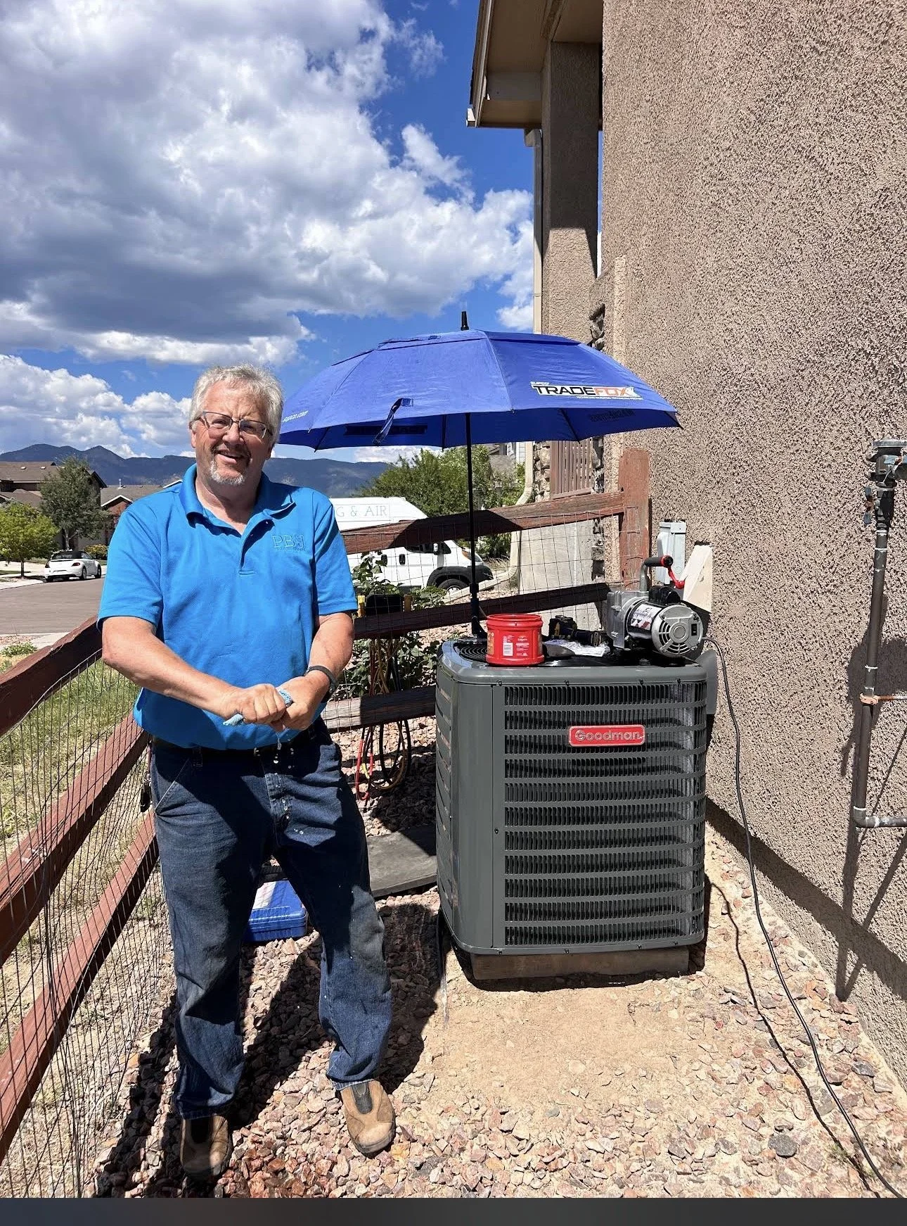A man wearing blue work clothes standing outdoors next to a Goodman HVAC unit, with a large blue umbrella, a power tool, and a red container on top of the unit, against a building and mountainous landscape under partly cloudy skies.