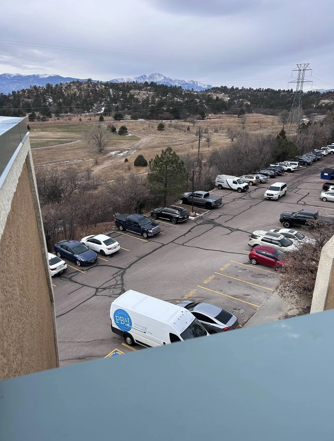 Parking lot with several cars and trucks, mostly white, black, and gray, surrounded by trees and open fields, hills, and mountains in the background under a cloudy sky.