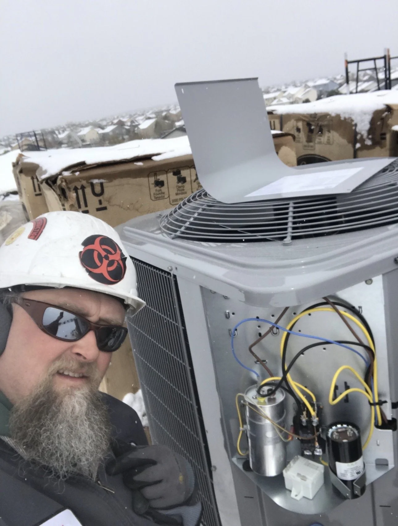 A man wearing a white safety helmet with a biohazard symbol, dark sunglasses, and a black jacket taking a selfie outdoors in snowy weather. Behind him is an open air conditioning unit with visible wires and components, and a partly cloudy sky.