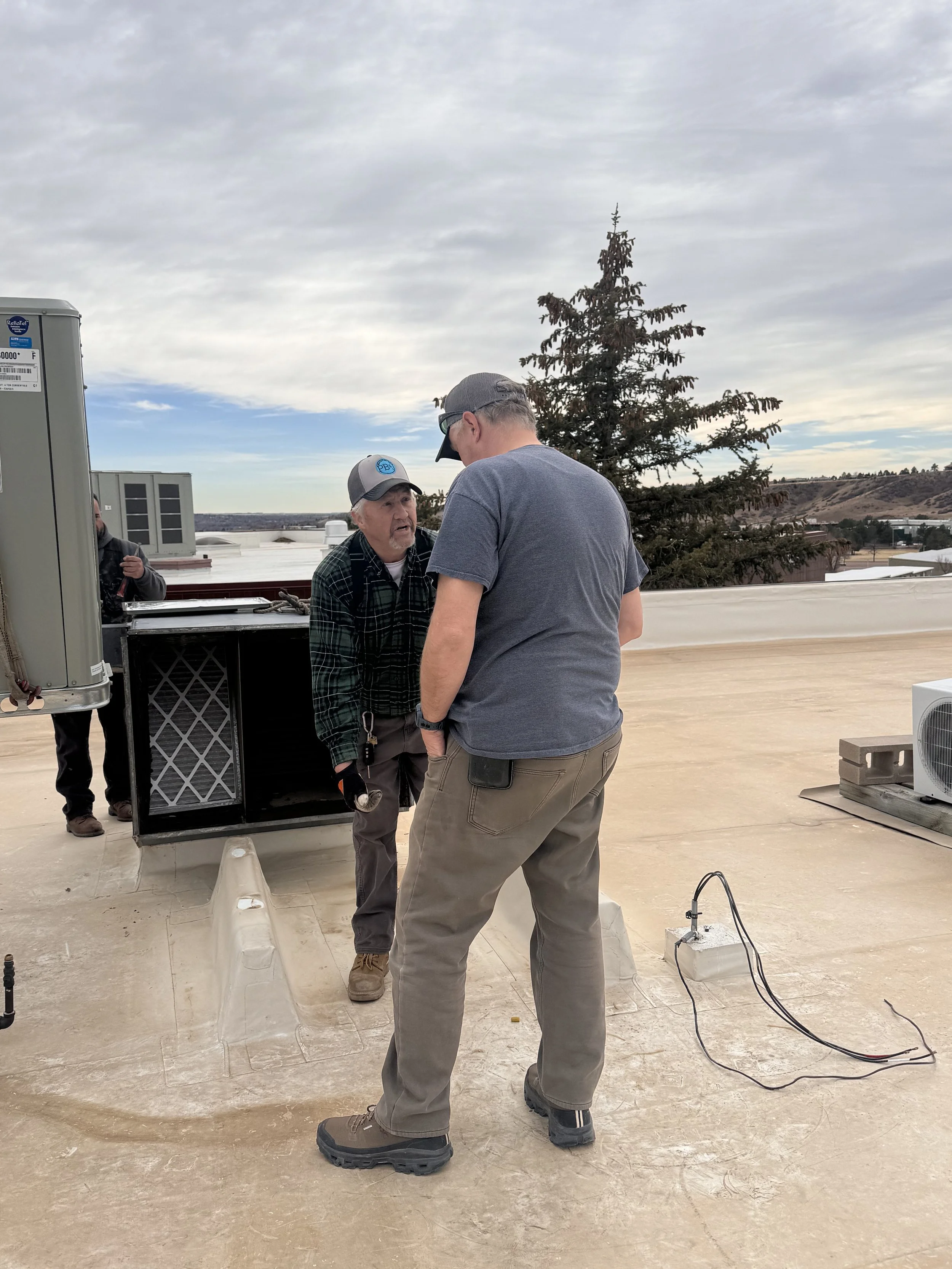 Two men standing on a rooftop, engaged in conversation. One man is facing away from the camera, wearing gray pants and a blue shirt. The other man is facing the camera, wearing a black jacket, a gray cap, and talking animatedly. There is a large elec