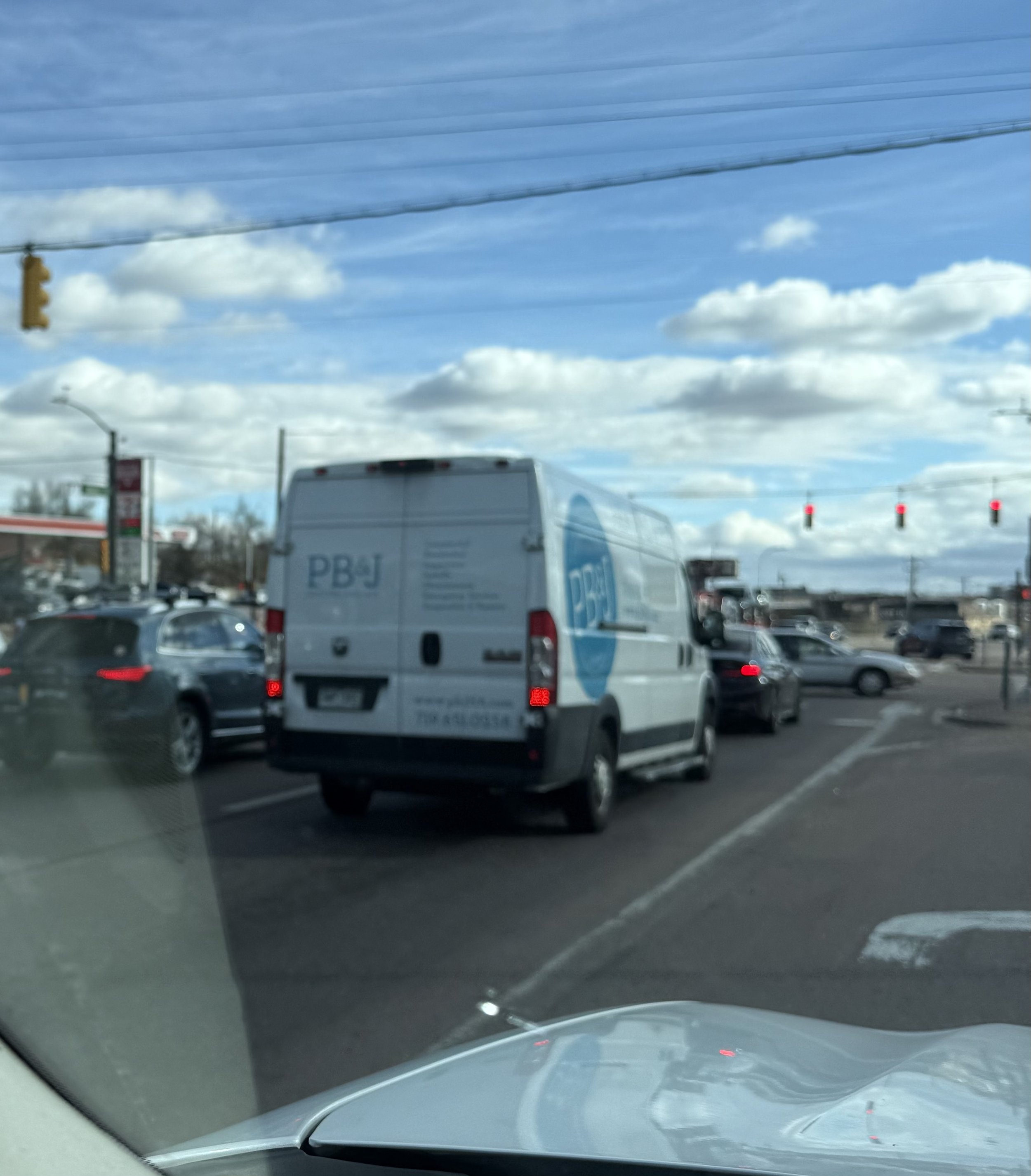 Traffic lights red, vehicles including a white van and cars on the road, blue sky with scattered clouds, power lines crossing the sky.
