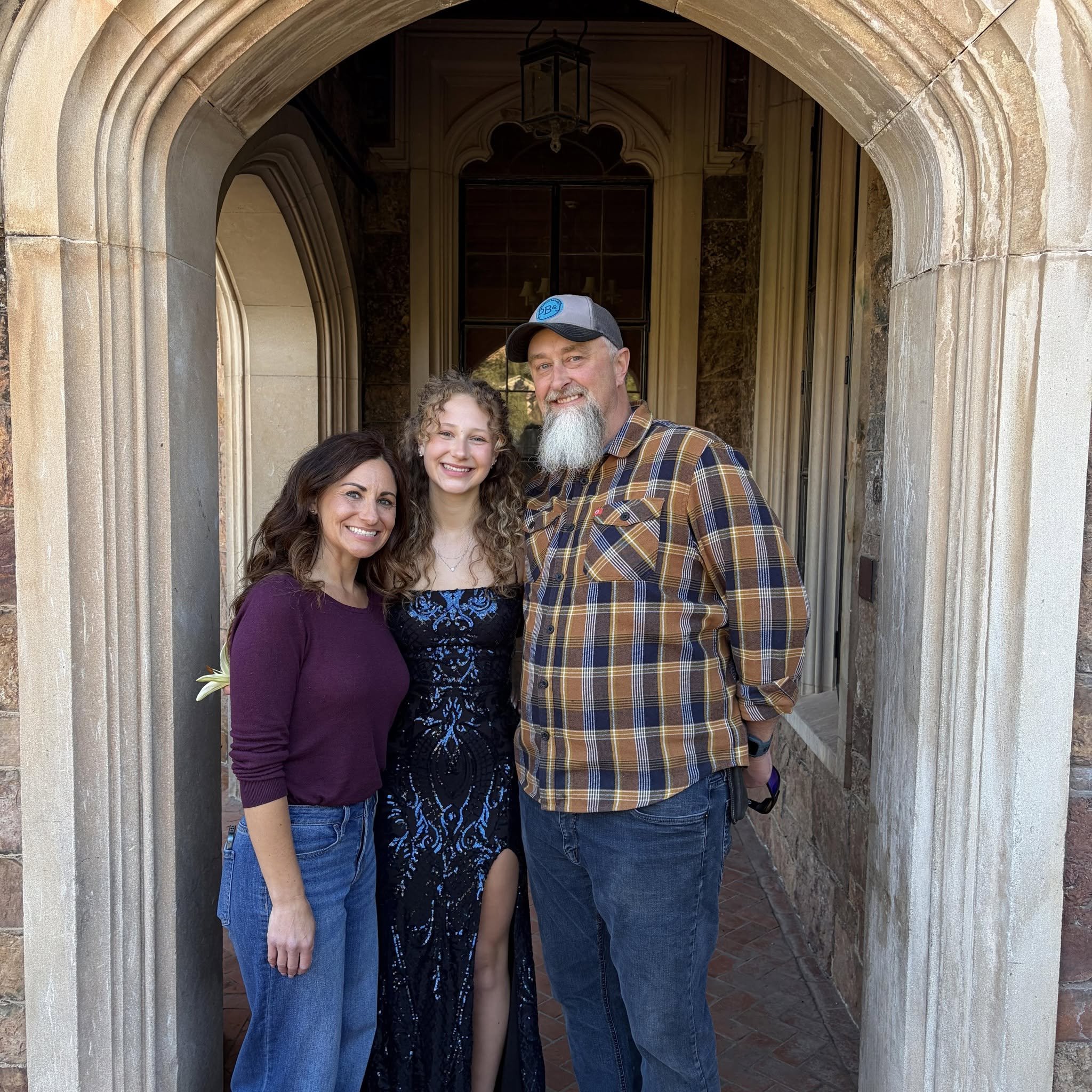A group of four smiling people, three women and one man, standing close together under an arched stone doorway. The man is wearing a baseball cap and a plaid shirt. The women are dressed in casual clothing, with one in a purple top and another in a black and blue dress. The setting appears to be outside a building with stonework and a window in the background.