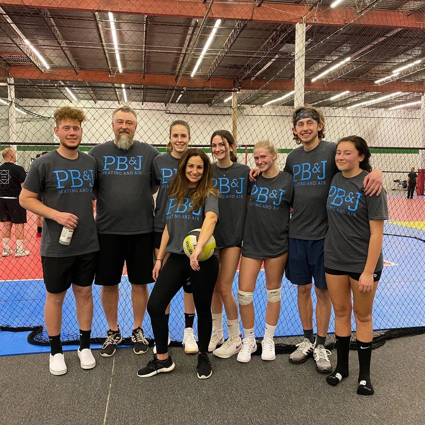 A group of seven people in matching gray shirts with the logo 'PB&J Heating and Air' posing together in an indoor sports facility with a volleyball net and court in the background.