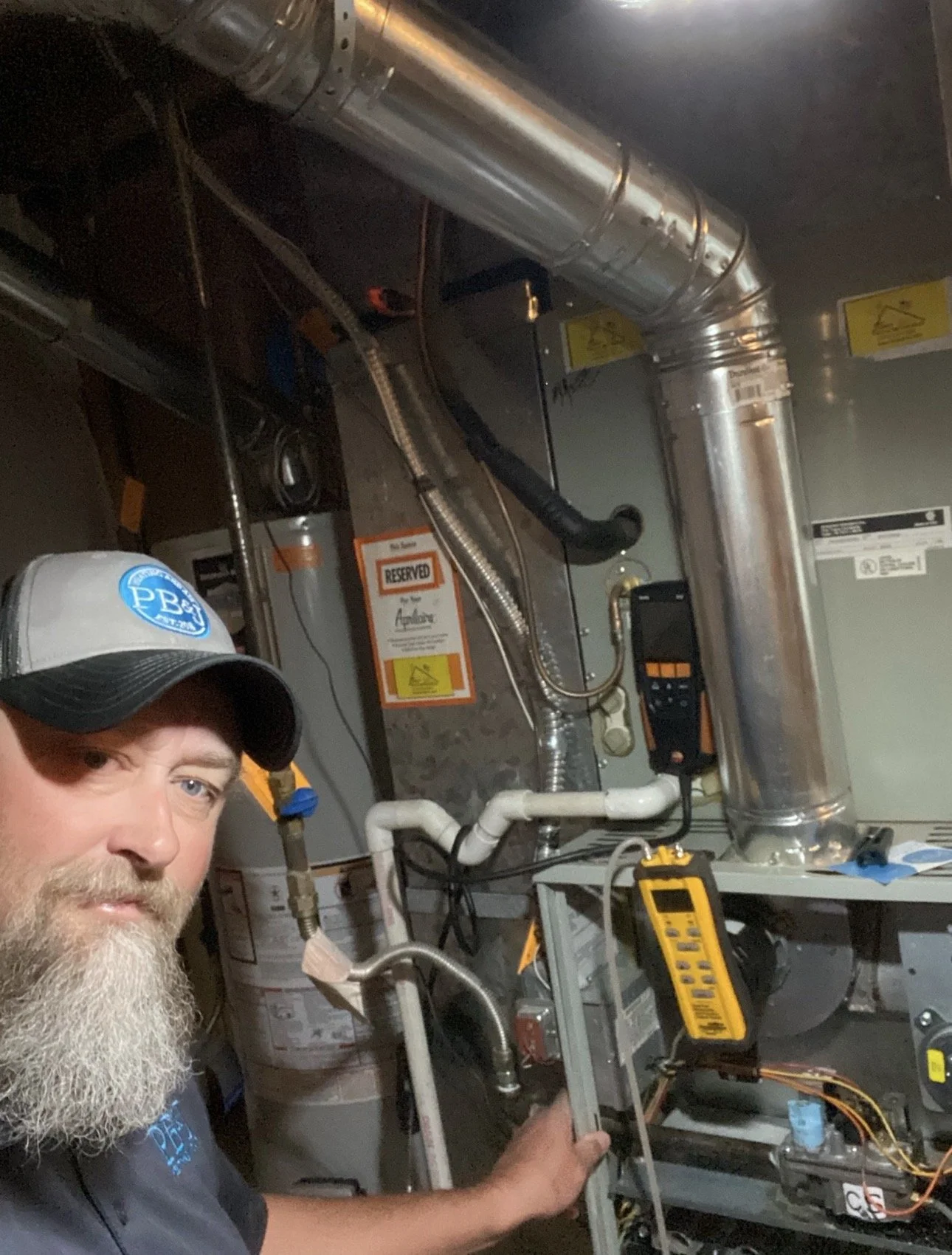 A man with a beard wearing a baseball cap and black work shirt operates HVAC equipment in a utility room, with various pipes, wires, and a multimeter visible.