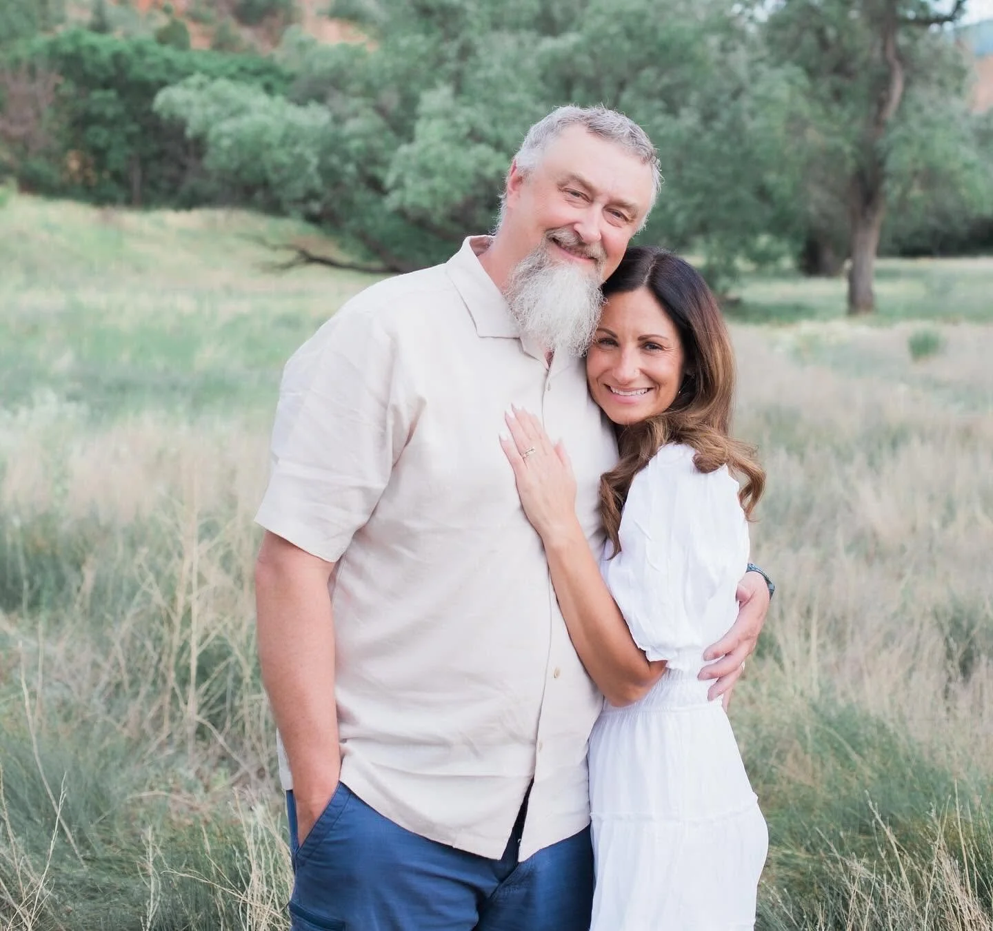A smiling man with gray hair and beard, wearing a beige shirt, standing outdoors with a woman who has long brown hair, wearing a white dress. They are hugging in a park with trees and grass in the background.