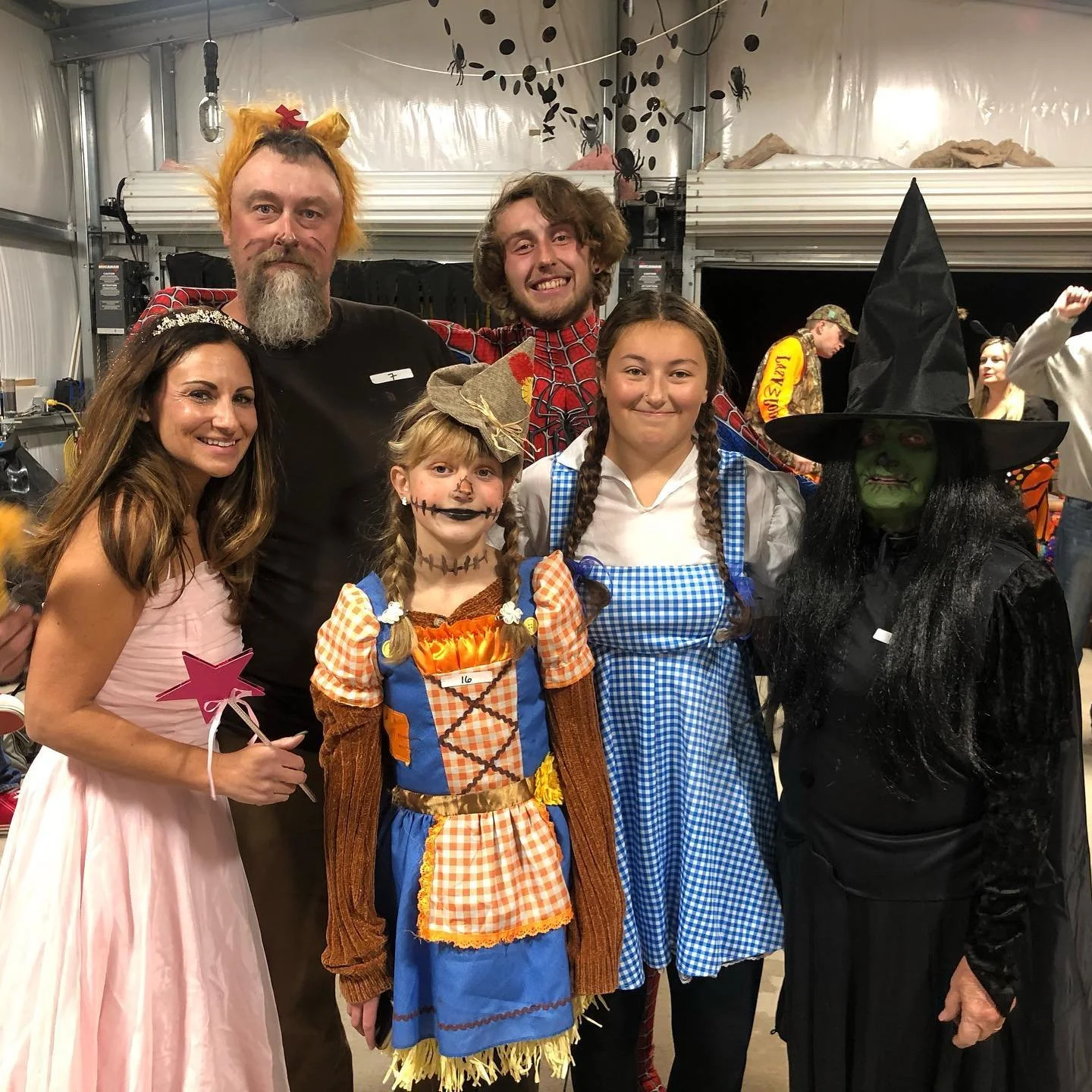 Group of six people in Halloween costumes inside a decorated event space. From left to right: a woman in a pink dress with a star wand; a man with orange hair, beard, and cat ears; a young girl dressed as a scarecrow; a man in a Spider-Man costume with a red and blue pattern; a girl dressed as Dorothy from The Wizard of Oz in a blue gingham dress; and a woman as a witch with a green face, black dress, and witch hat. Halloween decorations like black spider webs and hanging spiders in the background.