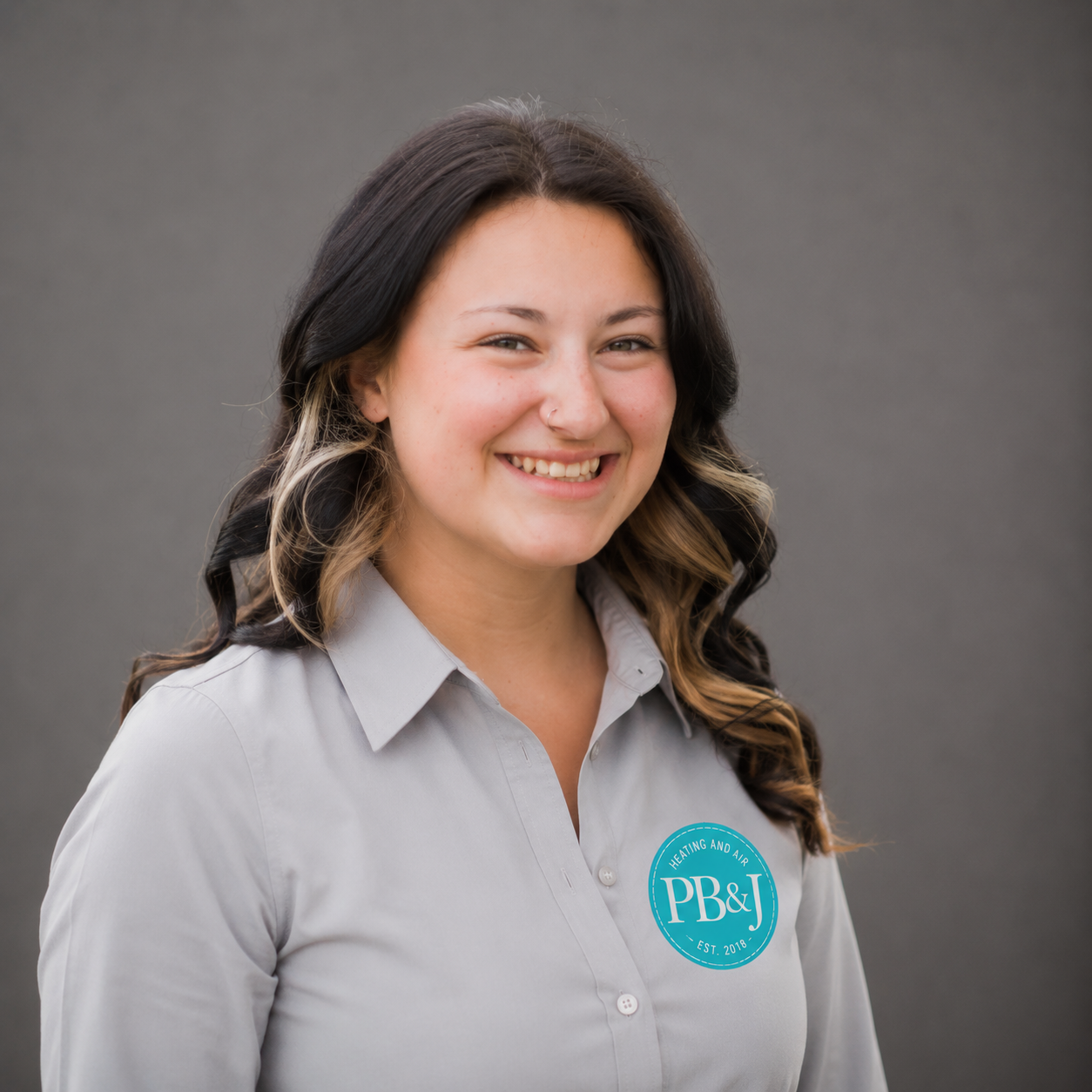 A young woman with dark and blonde wavy hair, wearing a light gray button-up shirt with a logo reading 'PB&J Heating and Air Est. 2018.' She is smiling in front of a neutral gray background.