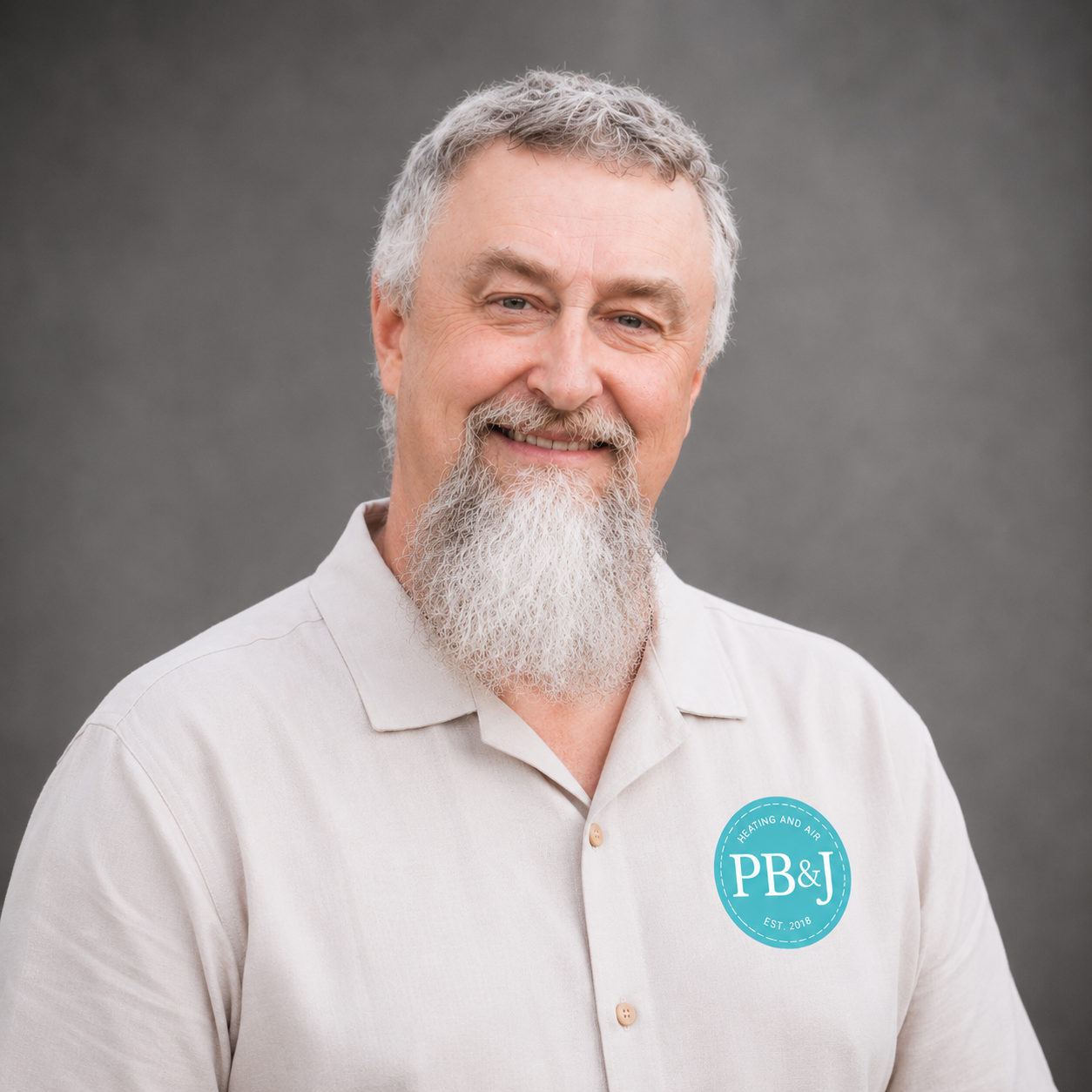 A smiling middle-aged man with gray hair and a full beard wearing a light-colored button-up shirt with a logo that says PB&J Heating and Air.