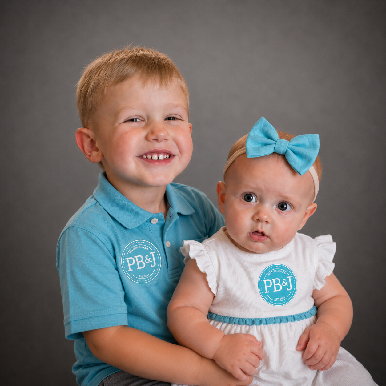 A young boy with red hair, wearing a blue polo shirt, smiling and holding a baby girl. The baby has a blue bow headband, wide eyes, and is wearing a white dress with blue accents. Both have a blue sticker on their shirts with the text 'PB&J' and are sitting against a gray background.