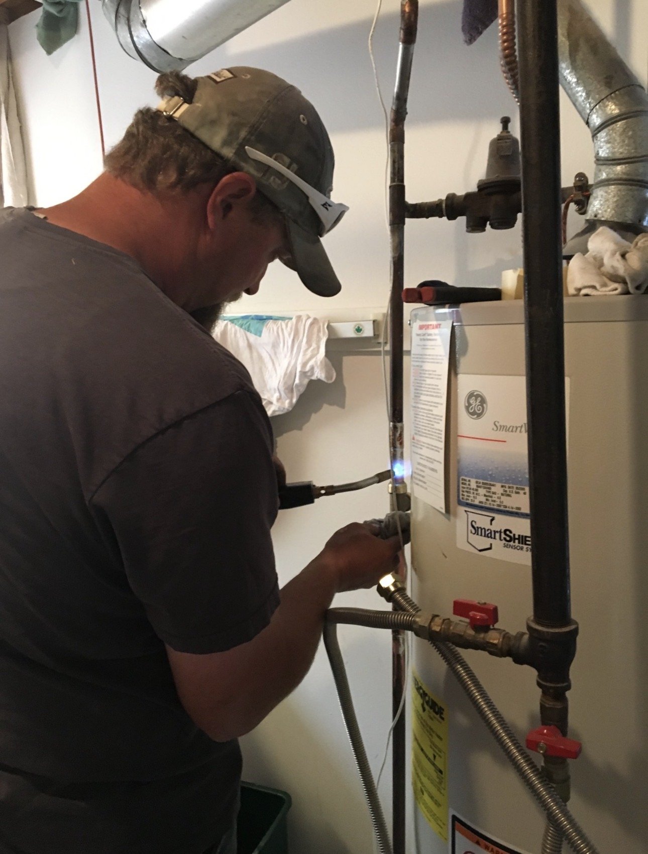 A man working on a water heater or boiler, wearing a cap and safety glasses, in a utility room with pipes and a towel hanging on the wall.