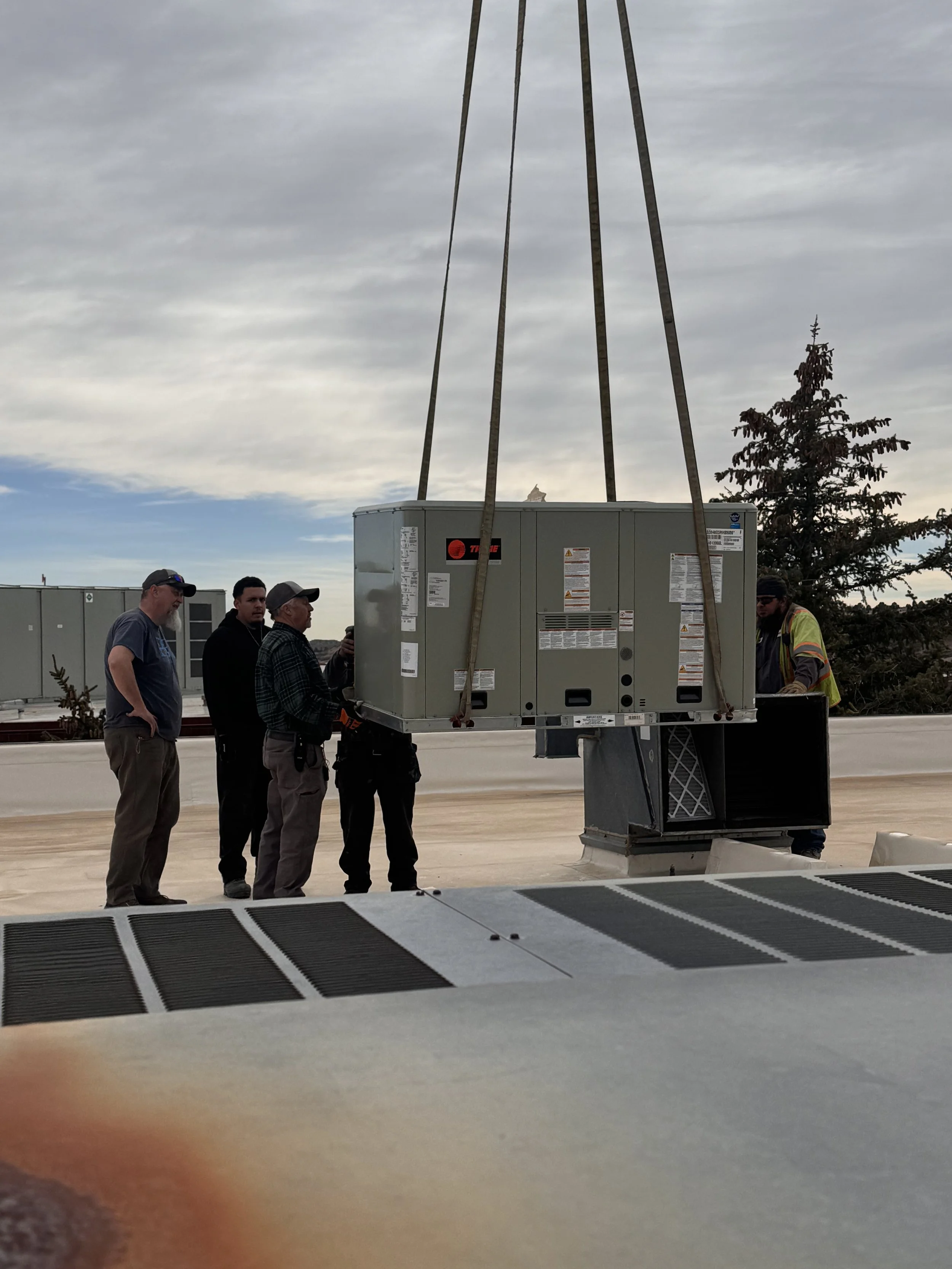 A group of five people are standing outside near a large electrical unit, with trees and a cloudy sky in the background. One person is wearing a high-visibility vest.