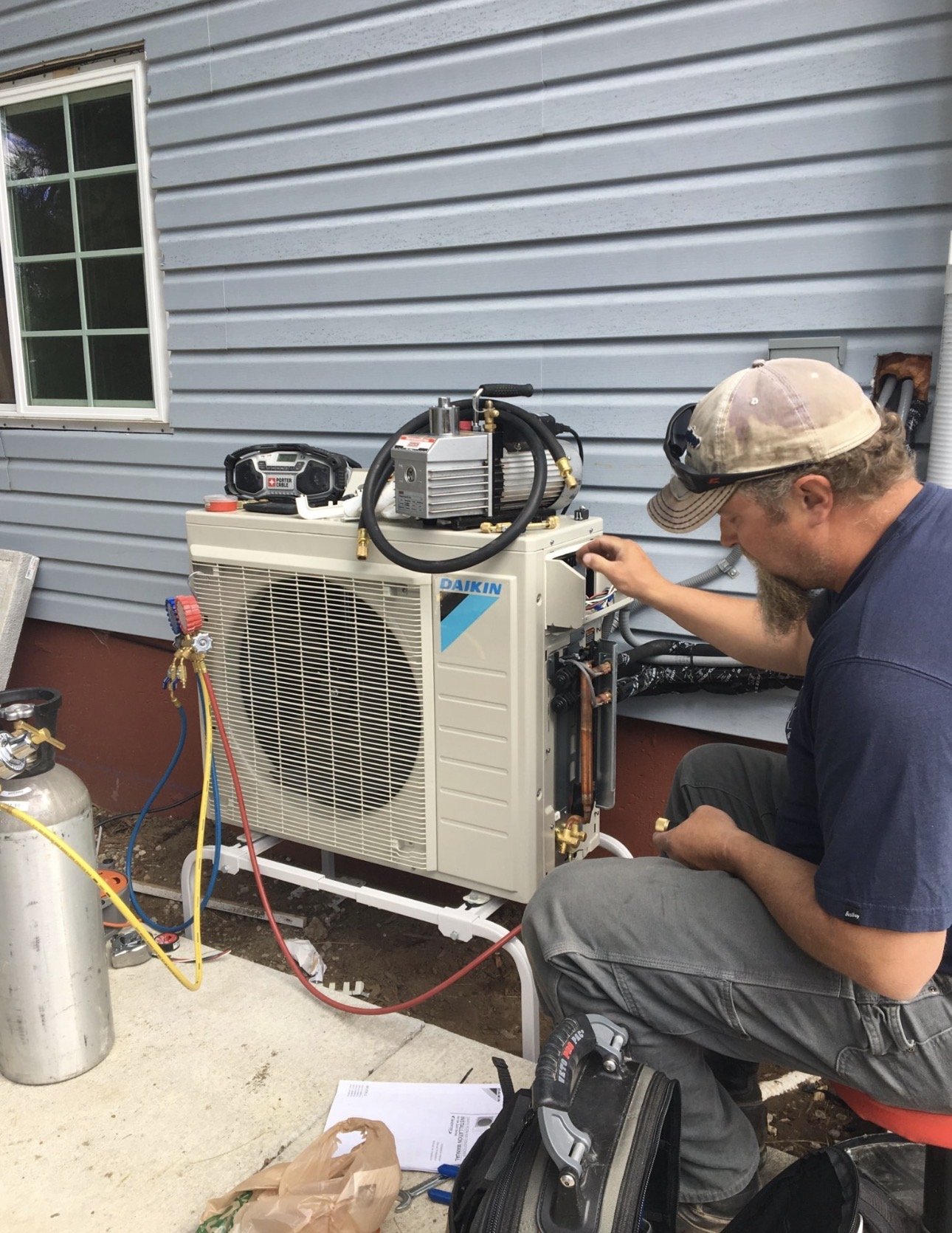 A technician is repairing or maintaining an outdoor air conditioning unit next to a house with light blue siding. The technician is wearing a beige cap and dark clothing, and is working with tools. There are various tools and equipment, including a p