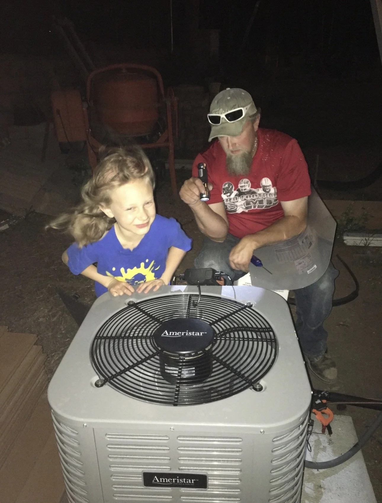 A young girl and a man working on an air conditioning unit outdoors at night. The girl is smiling and the man is wearing sunglasses and a hat.