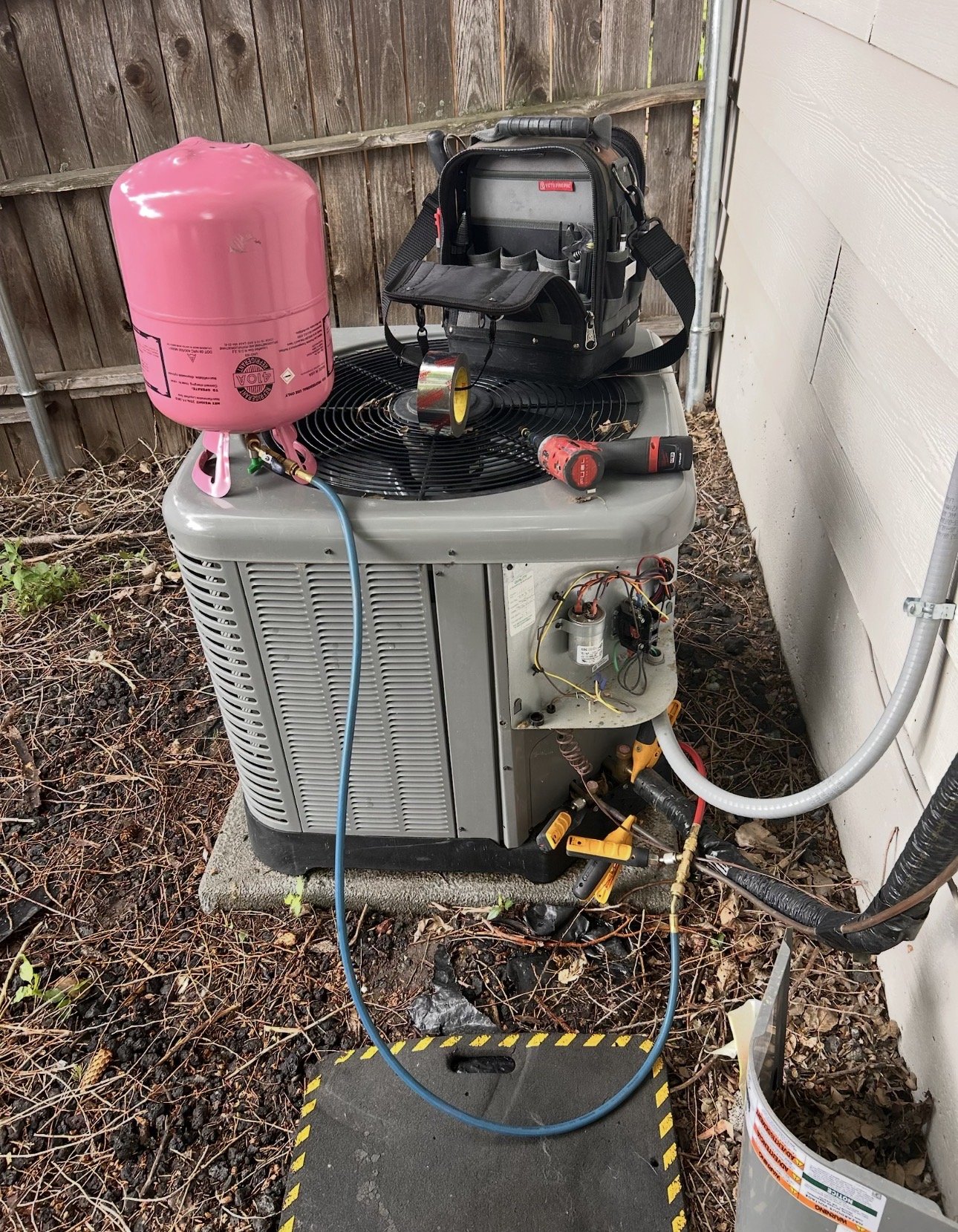 An outdoor HVAC unit with various tools and equipment on top, including a pink refrigerant tank connected with hoses, a black tool bag, a cordless drill, and some electrical wires, all placed on a concrete pad with a ground mat underneath.