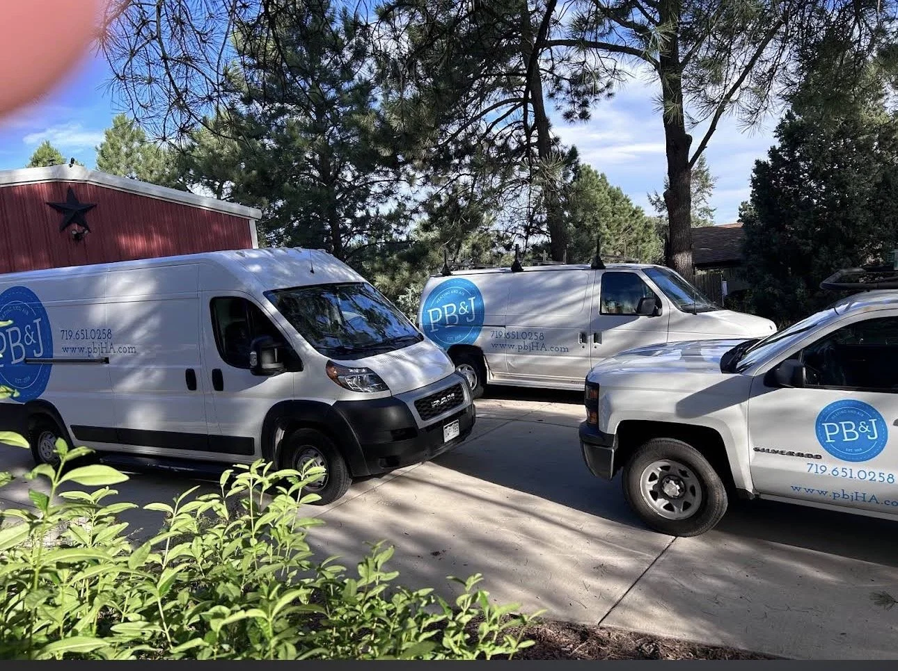 Three white service trucks with blue logos and contact information parked in a driveway, surrounded by trees and plants.