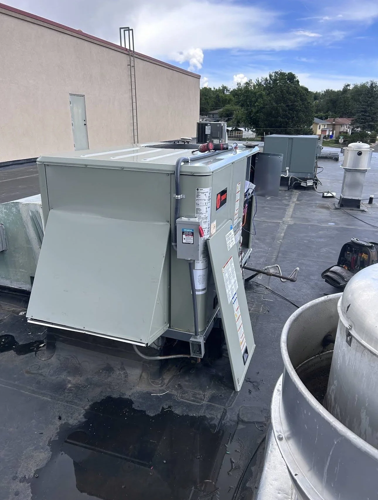 The image shows a rooftop with HVAC units and equipment, including a ladder attached to the building wall, a blue sky with clouds, and some trees in the background. There are tools and a bucket nearby.