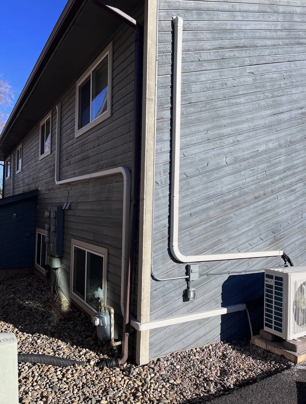 Back side of a house with an HVAC unit, electrical meters, and a plumbing system. Visible pipes and drain pipes run along the exterior wall, which is covered with grey horizontal siding. Gravel covers the ground below.