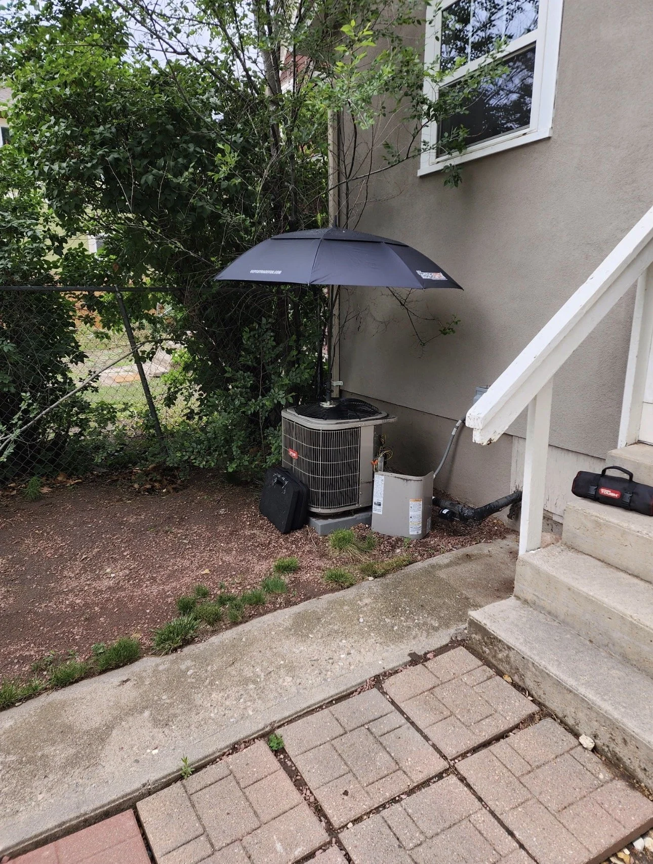 Air conditioning unit with a black umbrella attached to its top for shade, placed next to a house with stairs and a window, and surrounded by bushes and a fence.