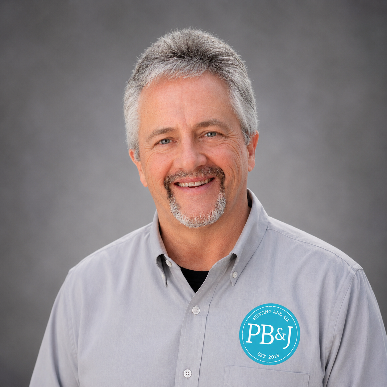 A smiling middle-aged man with gray hair and a beard, wearing a light gray button-down shirt with a P&J Heating and Air logo patch.