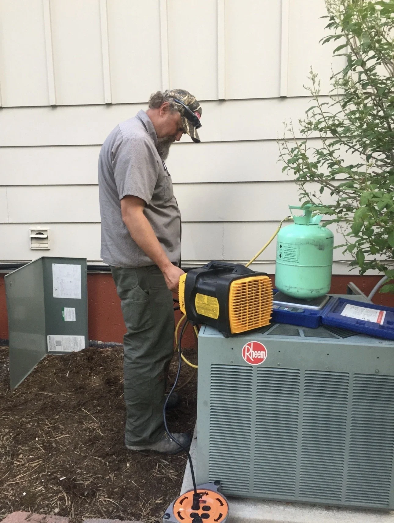 A man working on a heating, ventilation, and air conditioning (HVAC) system outside a house, wearing a cap and gray work clothes, with a fan and a green refrigerant tank nearby.