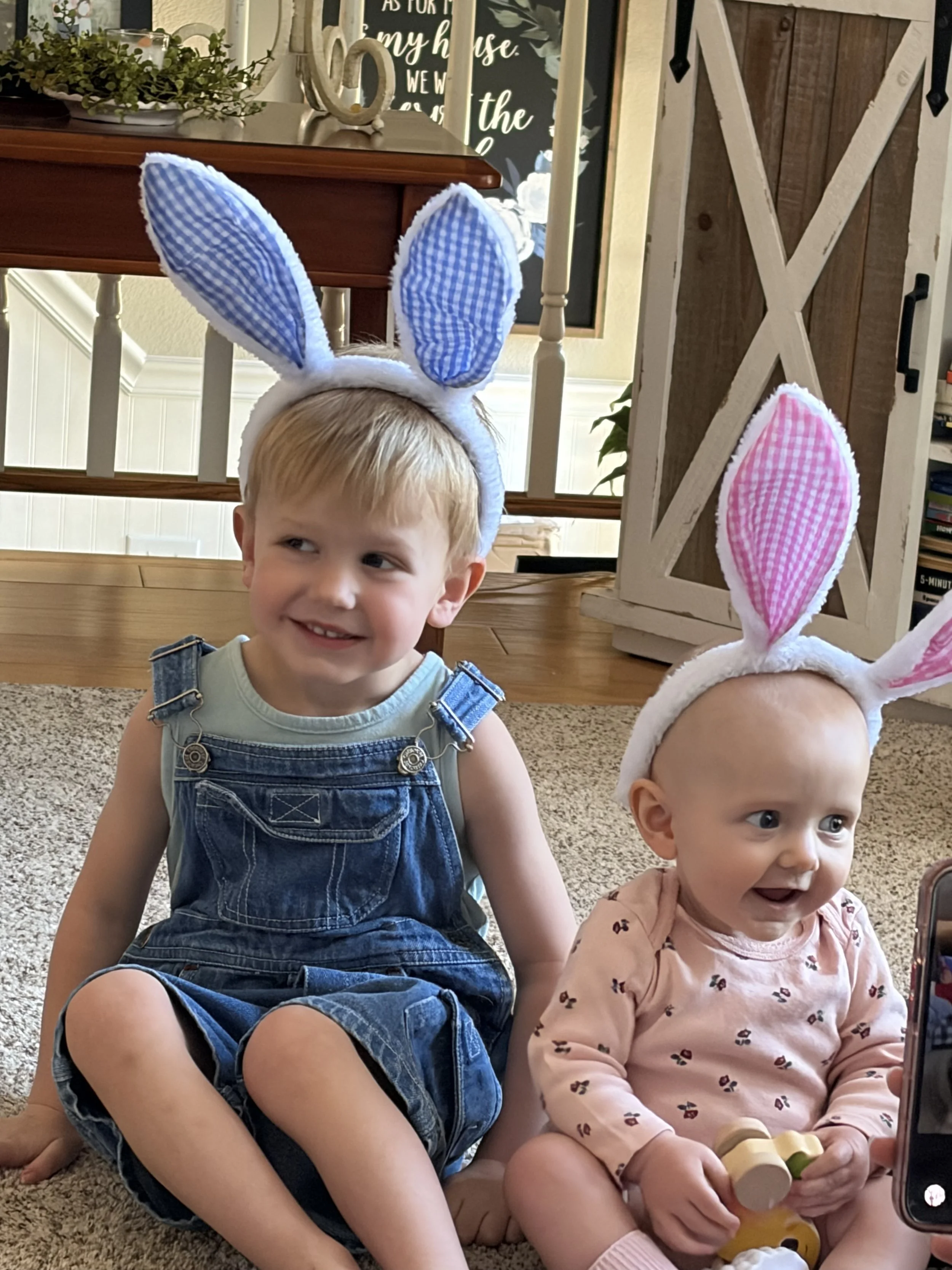 Two young children sitting on the floor, wearing bunny ear headbands with pink and blue gingham patterns. The boy has blonde hair, is smiling, and wearing denim overalls. The girl has light hair, wide eyes, and is holding a toy. A staircase, a wooden cabinet, and decorative items are visible in the background.