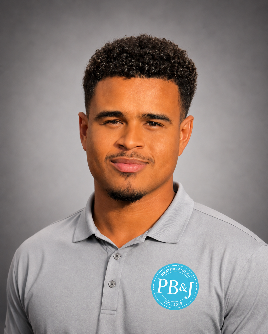 A young man with curly hair and a goatee wears a light gray polo shirt with a blue circular logo for PB&J Heating and Air. He is against a plain gray background.