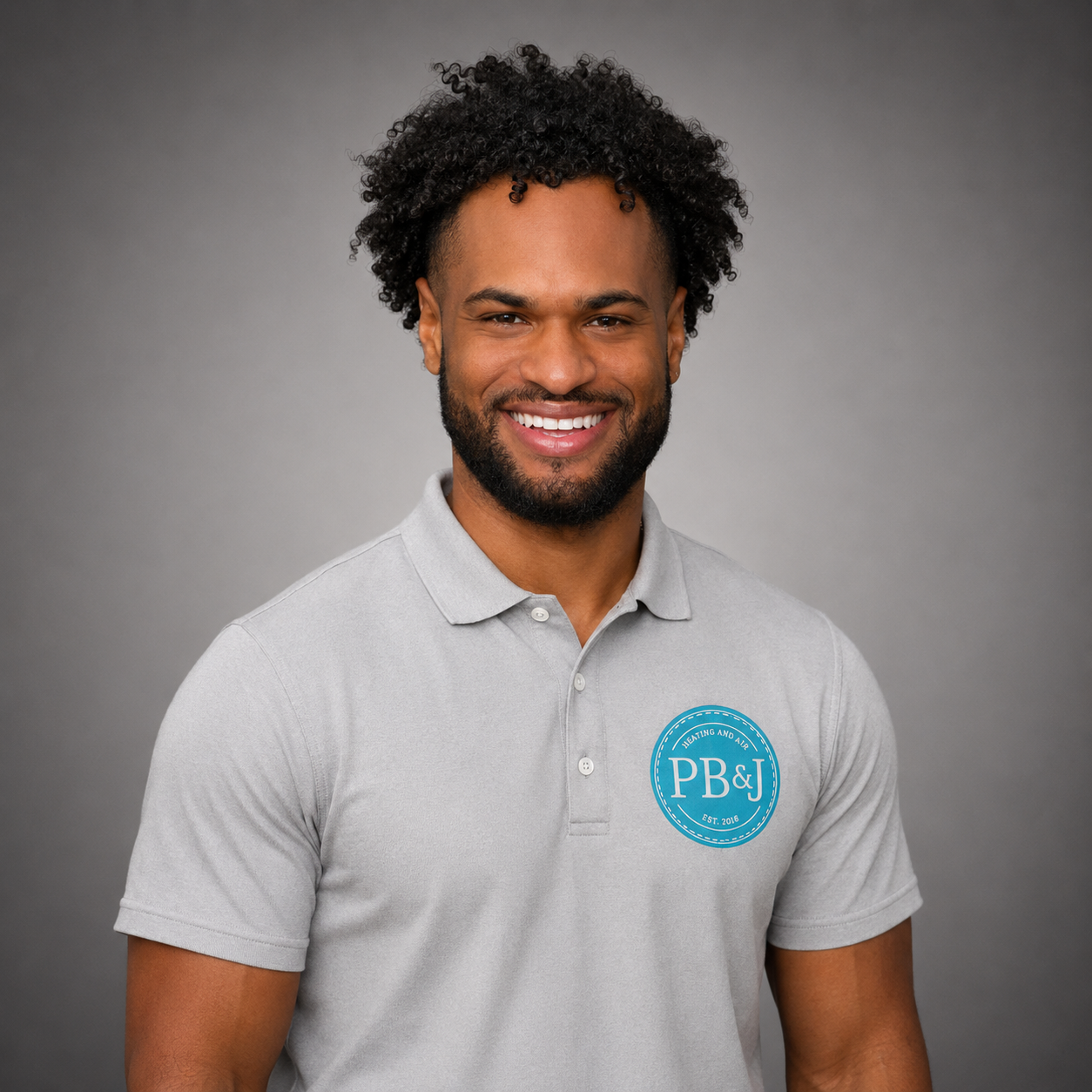 A smiling man with curly hair and a beard wearing a light gray polo shirt with a blue logo on the chest that reads 'PB&J' and 'Heating and Air' with a gray background.