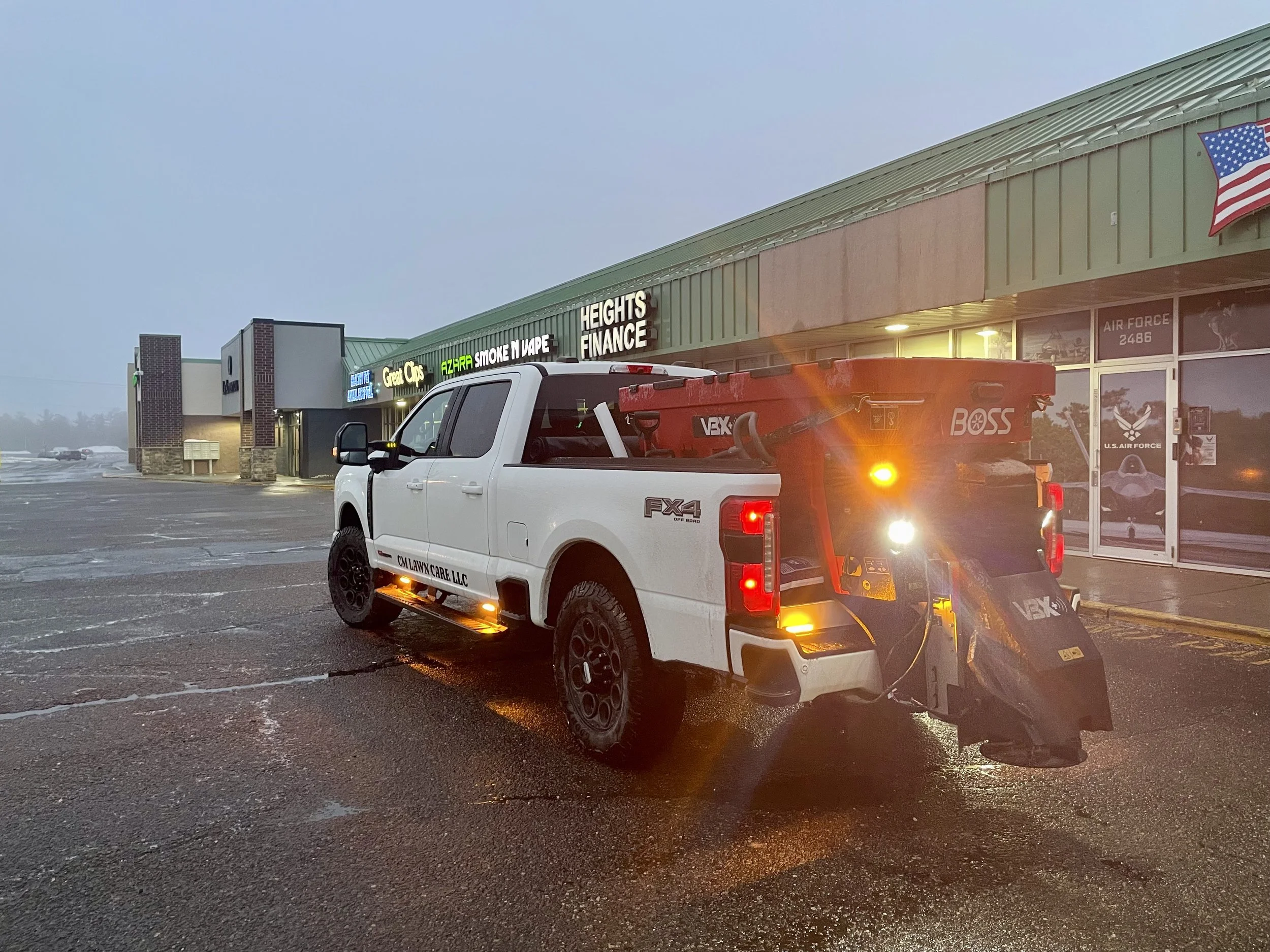 A parking lot outside a shopping center with stores, including AZARRA Smoke N Vape and Heights Finance, during evening or early morning.  CM Lawn Care LLC Winter Services salting