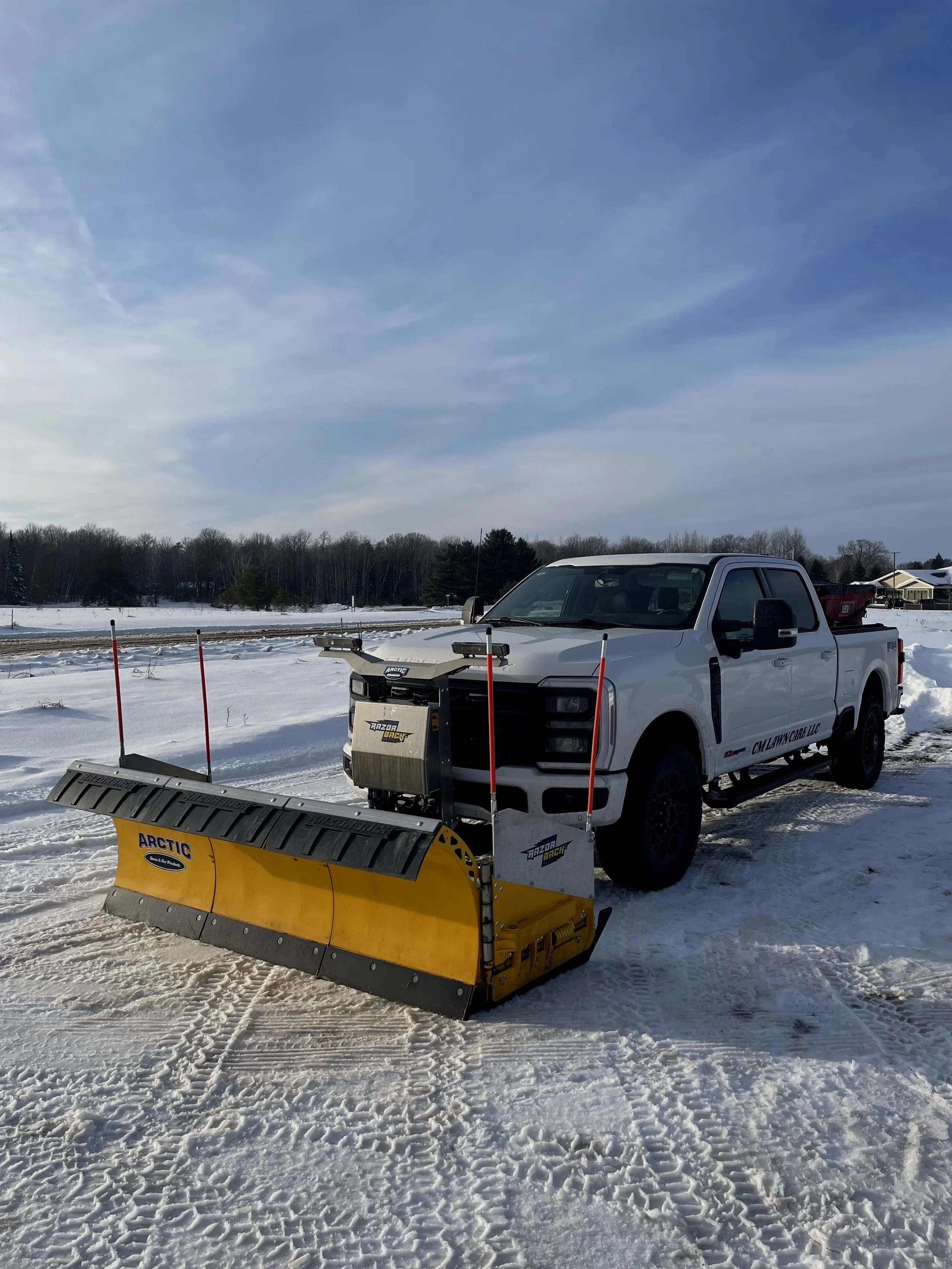 A white pickup truck with a yellow Arctic snowplow attached is parked on snow-covered ground in a rural area, with a background of trees and houses under a partly cloudy sky.