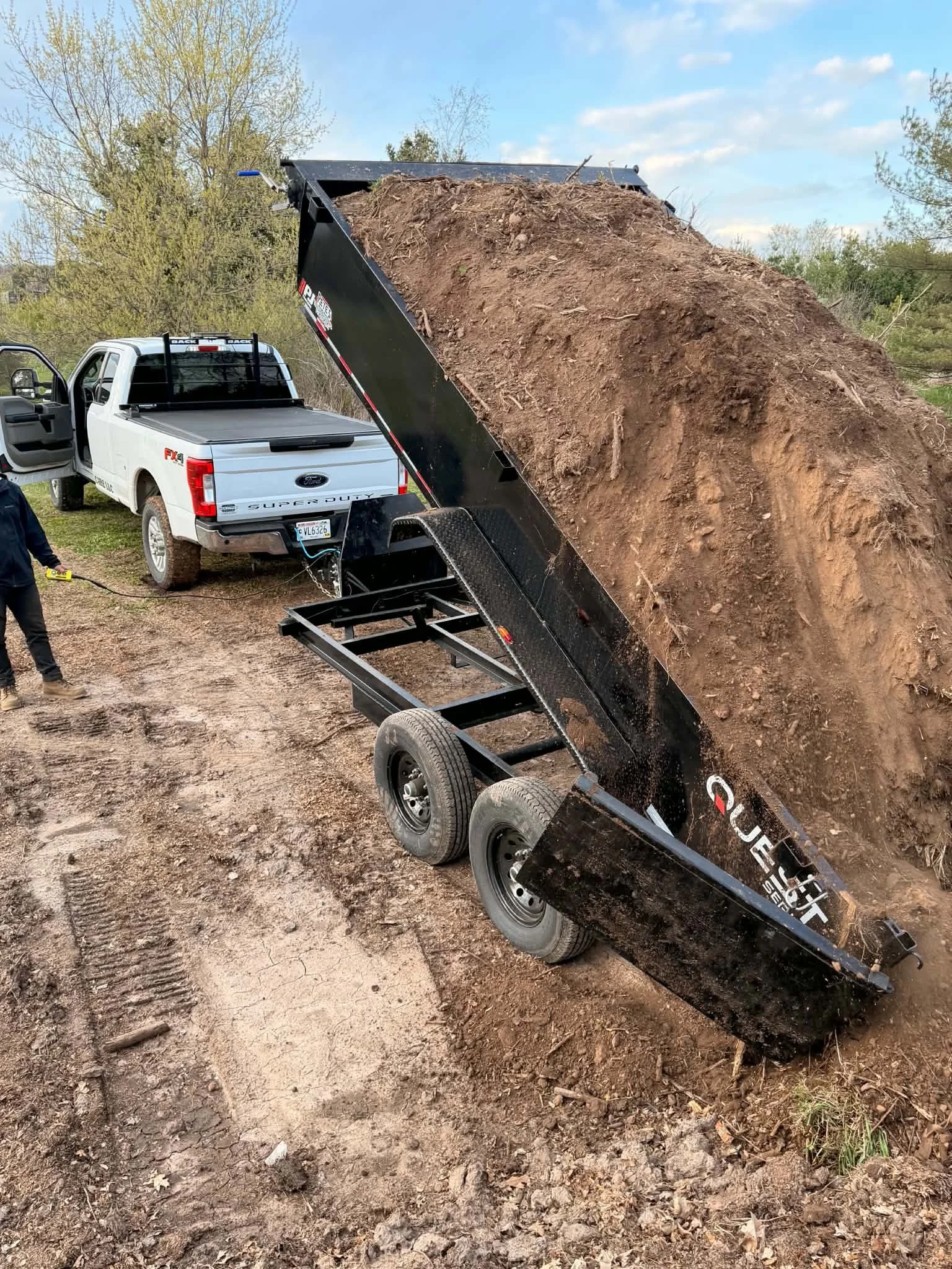A white Ford Super Duty pickup truck with a black dump trailer attached, dumping a load of dirt onto the ground outdoors, with trees and blue sky in the background. CM Lawn Care LLC
