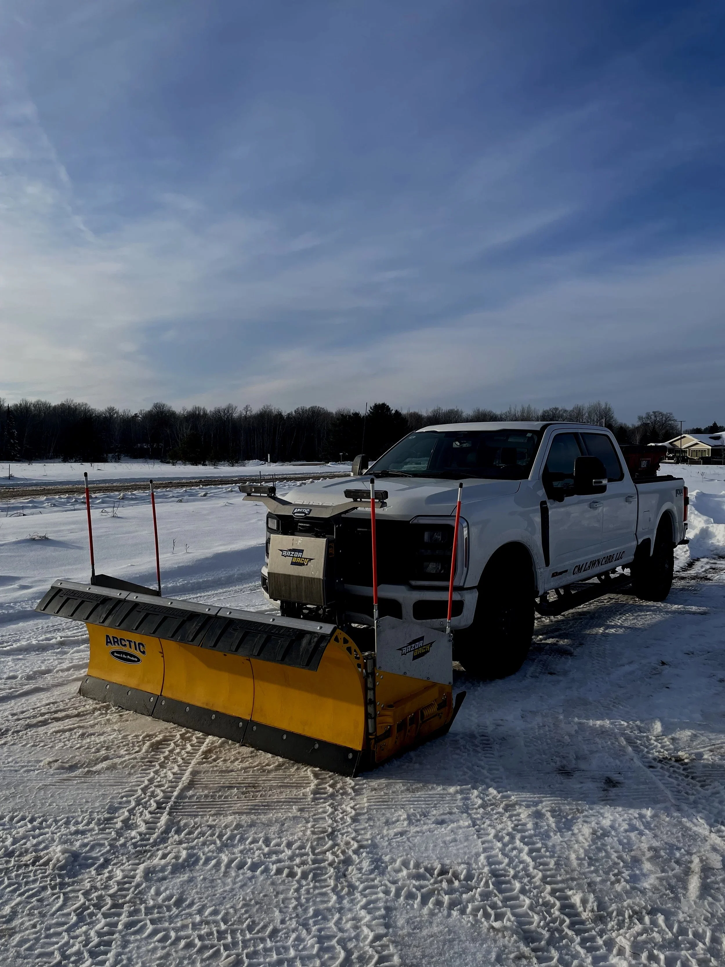 A white pickup truck equipped with a yellow Arctic snow plow attachment parked on snow-covered ground in a winter landscape with a cloudy blue sky and a tree line in the background.