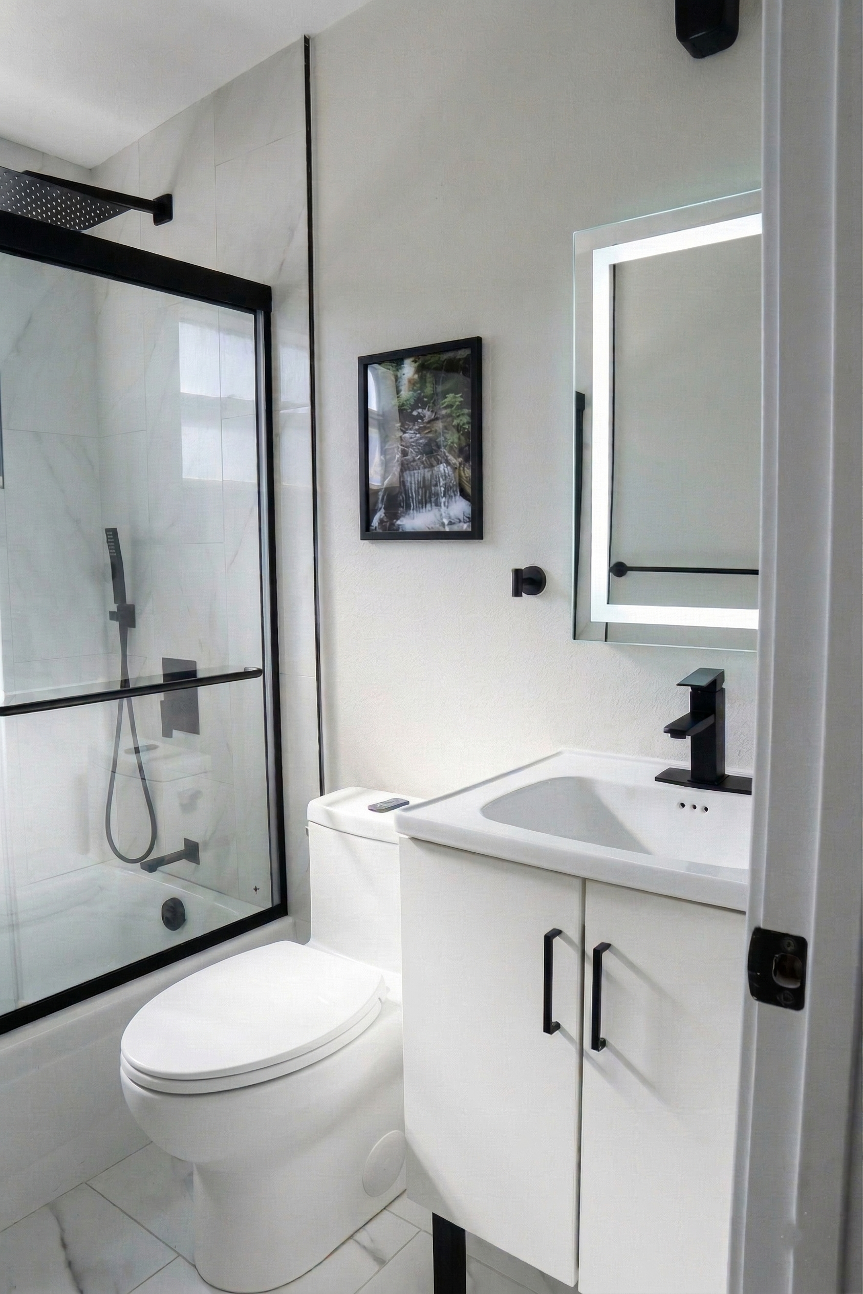 Modern bathroom with a walk-in shower with black fixtures and a glass door, a white toilet, a white vanity with black handles, a rectangular mirror with LED lighting, a framed nature photograph on the wall, and white marble tile flooring.