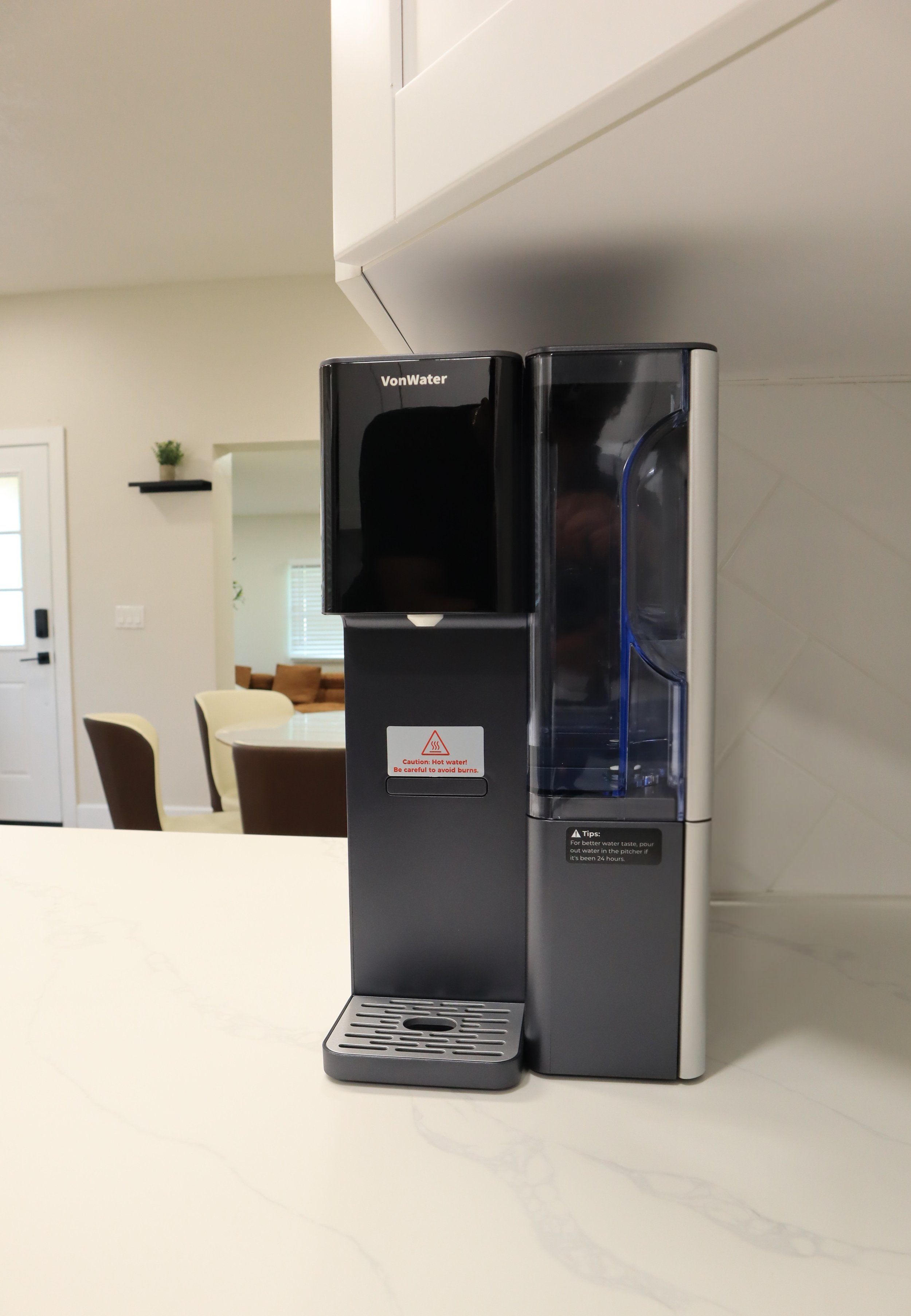 Black water dispenser on a white table in a modern kitchen with a living room in the background.