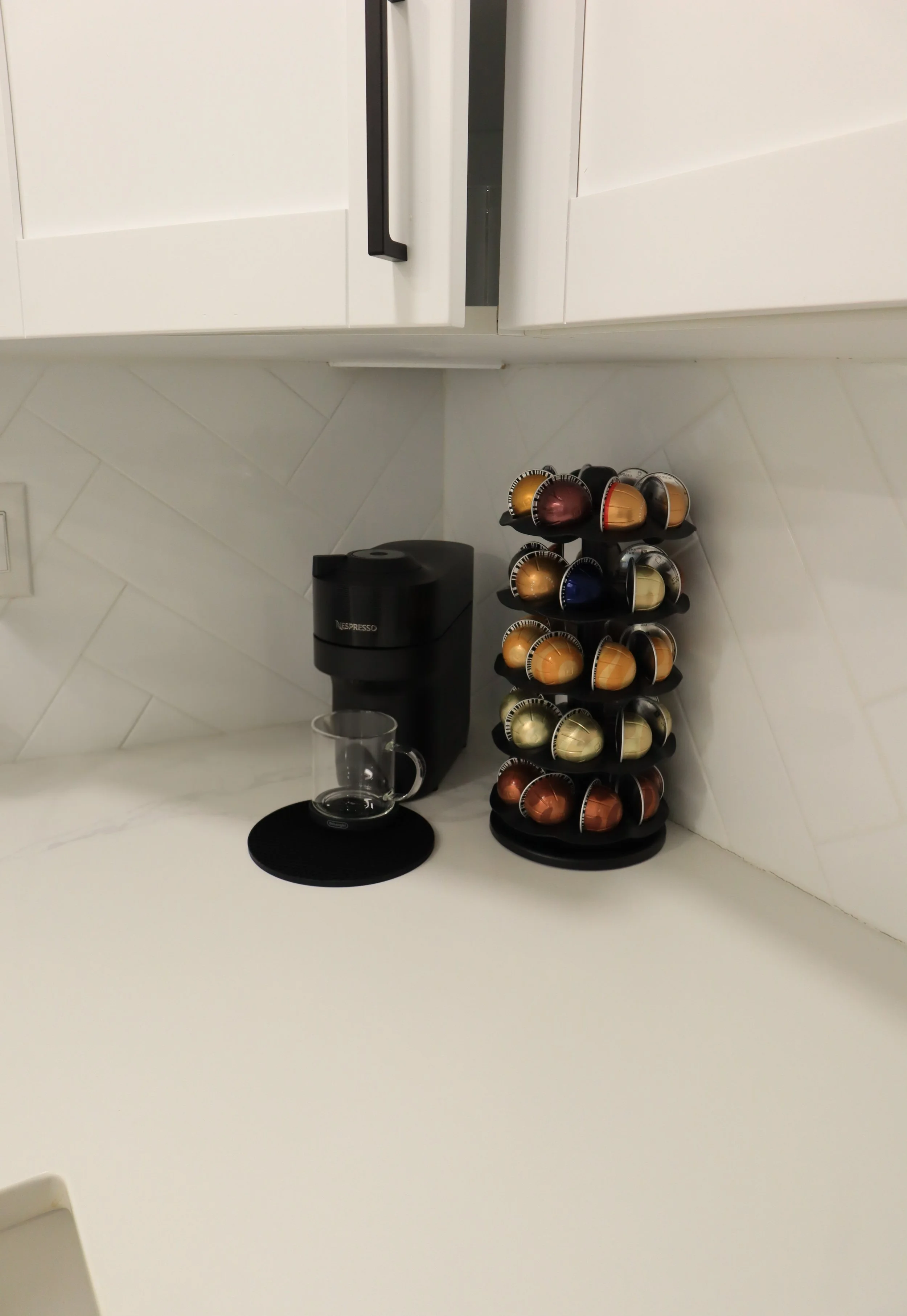 Kitchen countertop with a Nespresso coffee maker, a glass mug, a black mat, and a rotating capsule holder filled with various coffee capsules.
