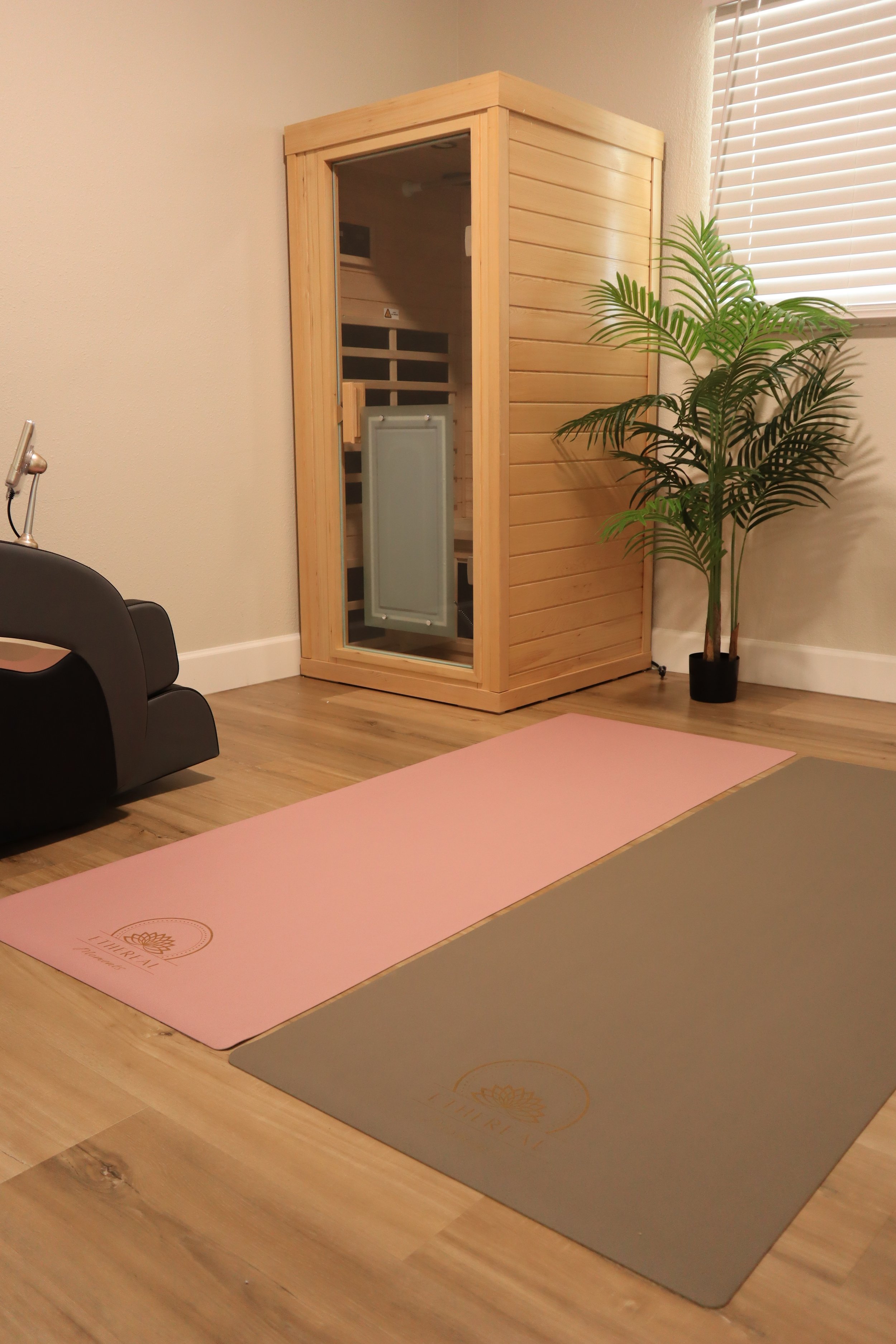 Interior of a yoga or meditation studio with two yoga mats, one pink and one gray, placed on a wooden floor. There is a wooden sauna or changing room with a glass door, a plant in a black pot, and a window with blinds on the right side.