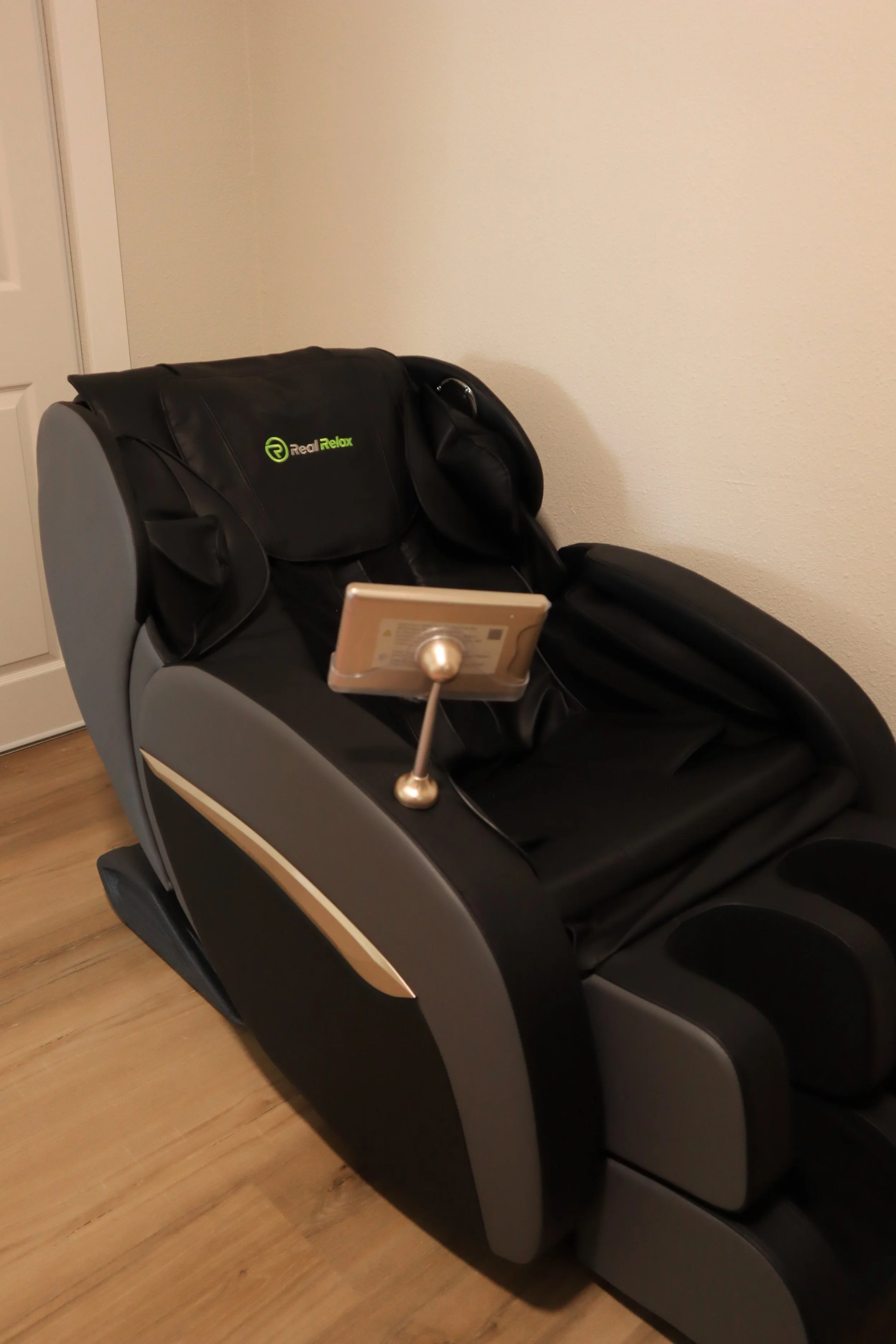 A black massage chair with a small electronic device attached to it, set against a beige wall on a wooden floor.